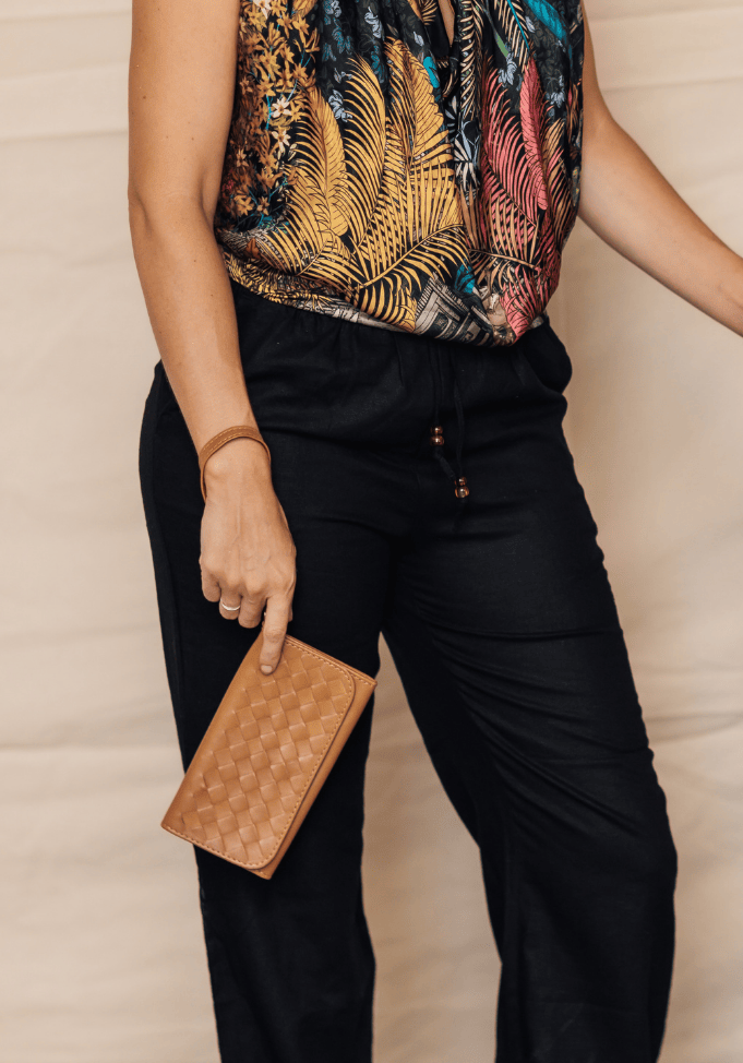 A lifestyle shot of a woman wearing a tropical print top and black pants, holding a brown woven clutch. She is standing next to another person in a beige dress holding a brown leather shoulder bag, against a neutral beige backdrop.