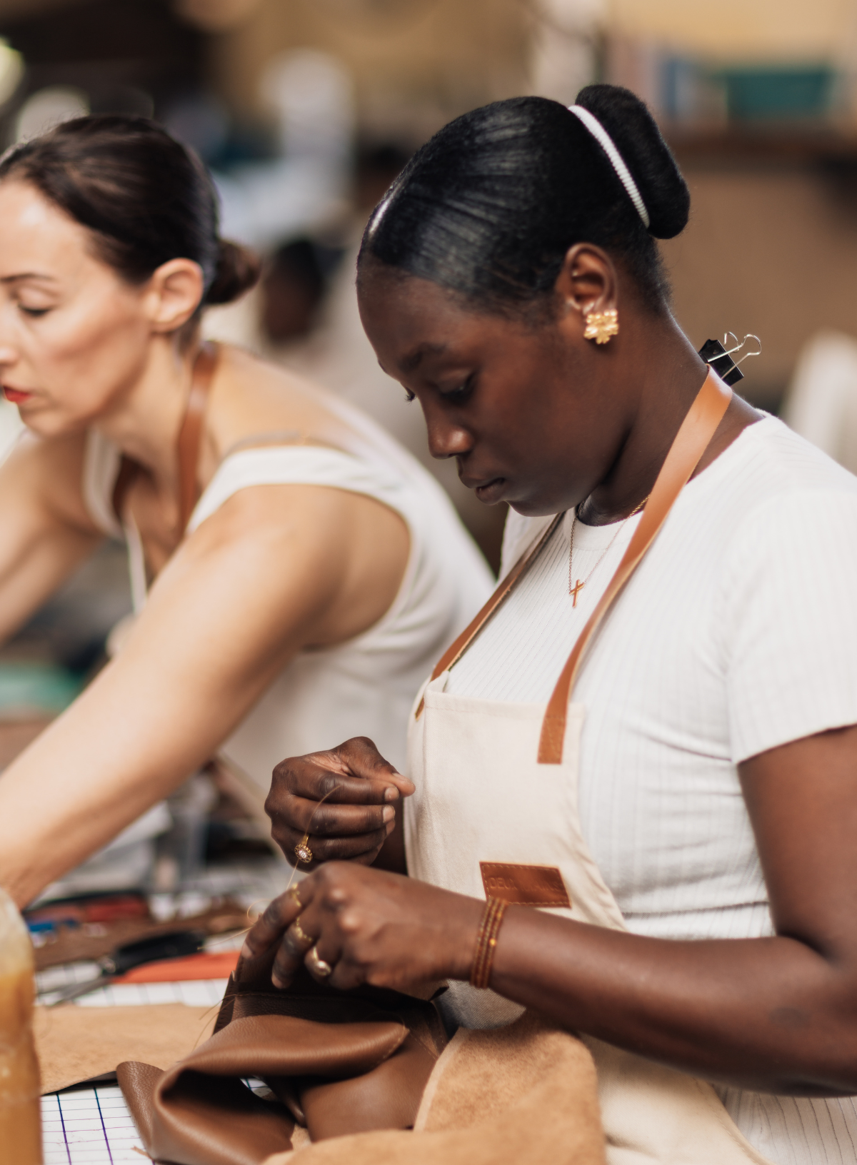 A woman in a white apron works at a leathercraft table, shaping a brown leather piece, with another woman working in the background in a warm, softly blurred workshop setting.