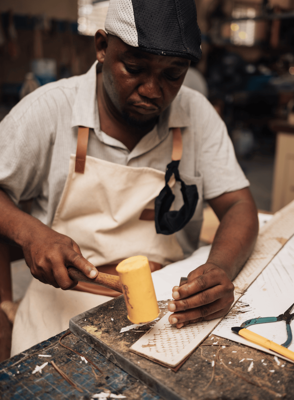 A craftsman wearing a beige apron and a black-and-white cap is shown in a workshop, using a yellow mallet to work on a piece of wood or metal on a workbench.