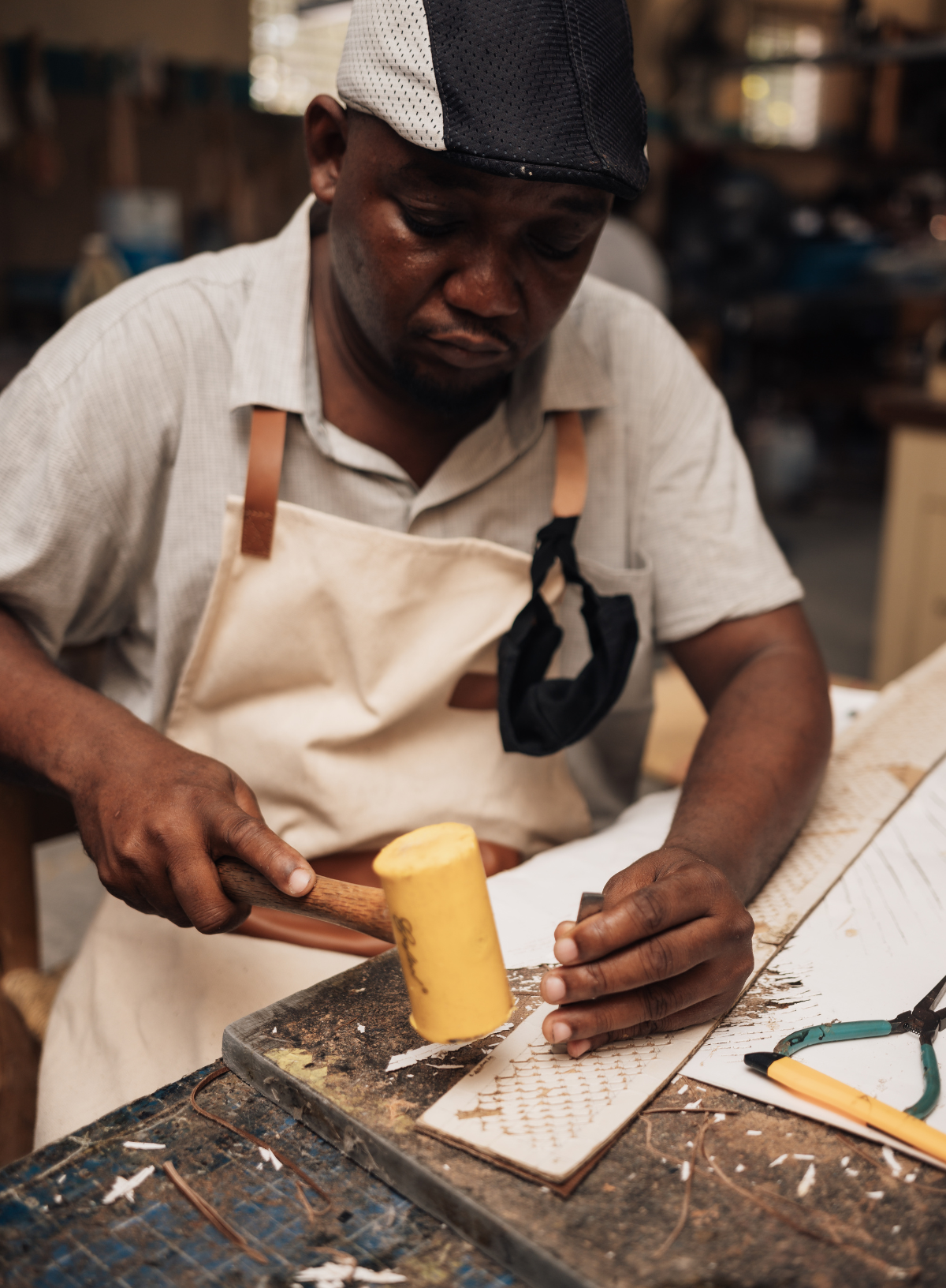 A craftsman wearing a beige apron and a black-and-white cap is shown in a workshop, using a yellow mallet to work on a piece of wood or metal on a workbench.