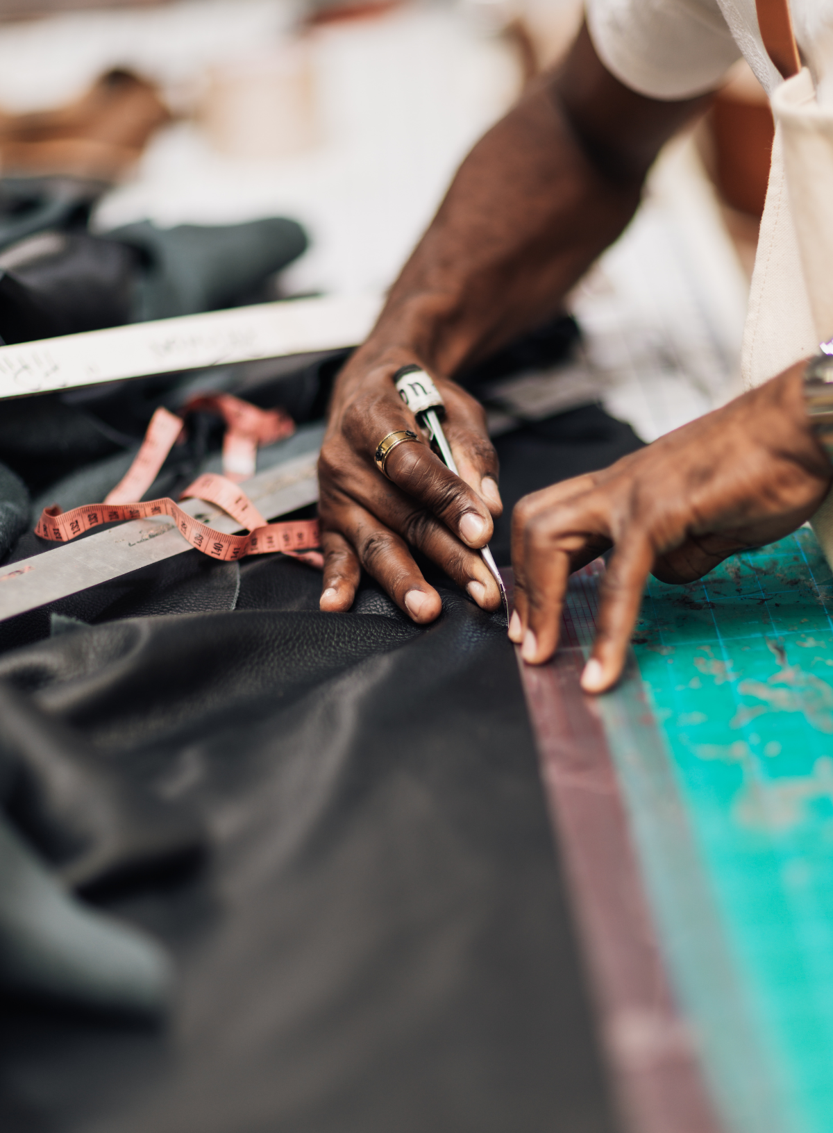 Close-up of a tailor's hands marking black fabric with a pencil, surrounded by measuring tools like a tape measure and ruler on a worktable.