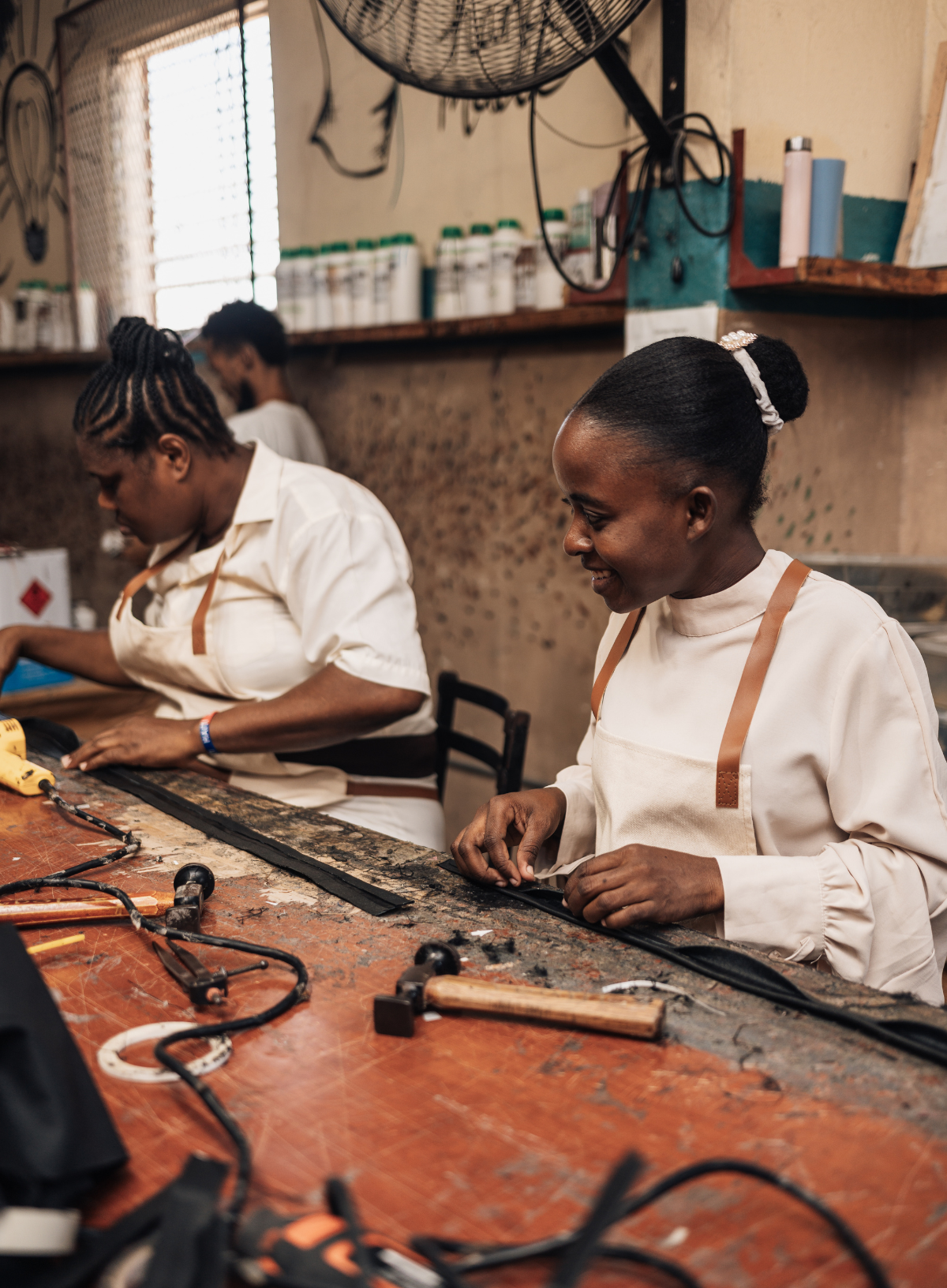 Two women working at a wooden craft table in a workshop, wearing matching white long-sleeve tops with brown shoulder straps.