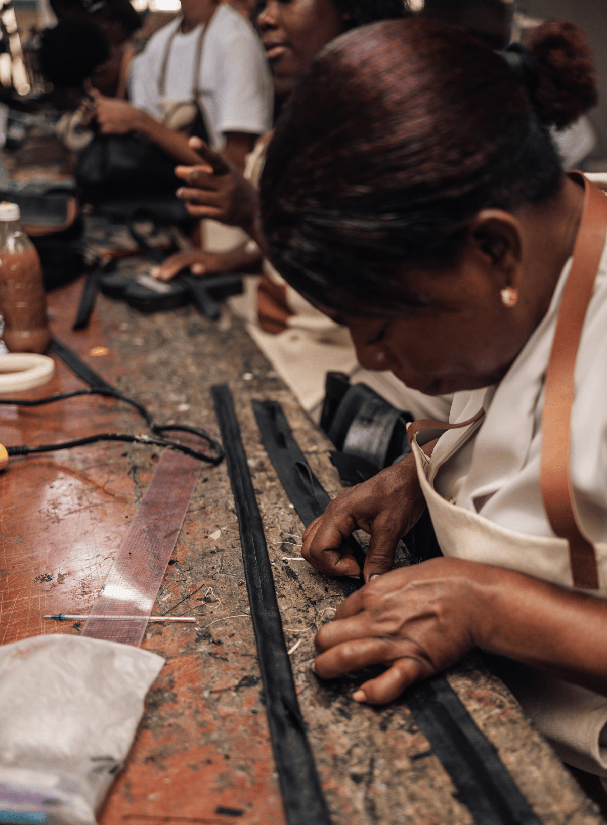 A close-up lifestyle shot of a woman working at a leathercraft table, carefully stitching black leather strips with a needle and thread. She is wearing a white apron, and other crafting tools and people are visible in the background of the workshop.