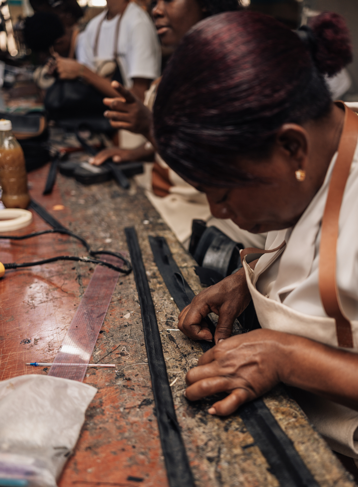 A close-up lifestyle shot of a woman working at a leathercraft table, carefully stitching black leather strips with a needle and thread. She is wearing a white apron, and other crafting tools and people are visible in the background of the workshop.