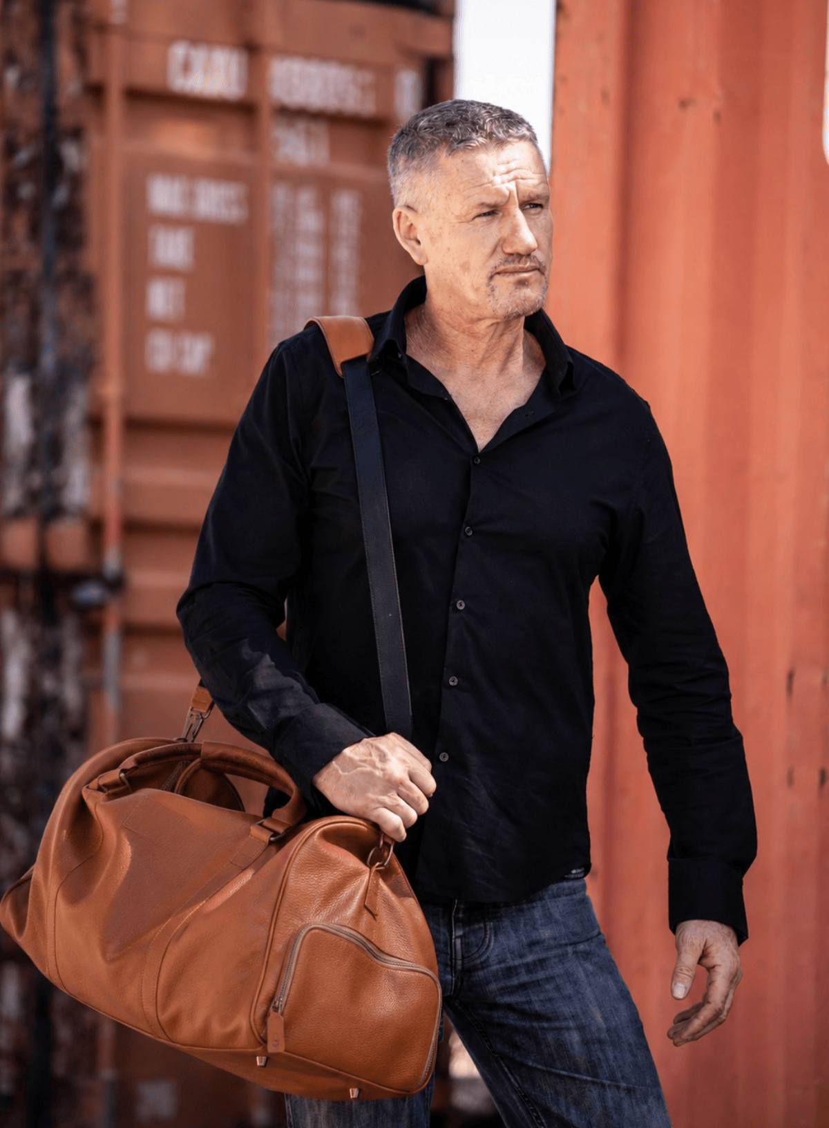 Man holding a brown leather bag against a red shipping container background