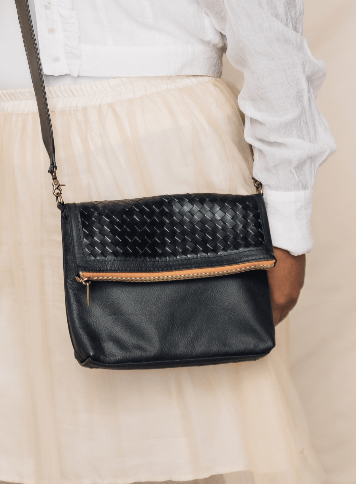A black faux-leather crossbody bag with a woven basket-texture flap and a tan zipper accent, shown worn over the shoulder of a person in a white blouse and cream skirt.