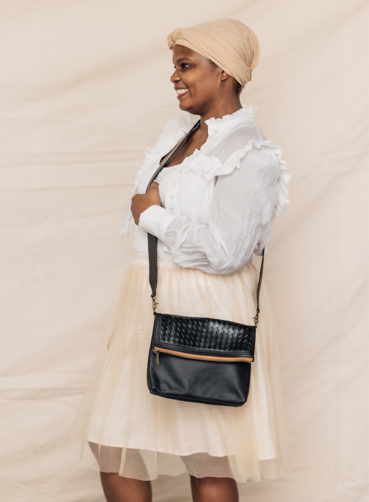 A woman wearing a beige headwrap, a white ruffled blouse, and a cream pleated skirt carries a black crossbody bag with a textured front and tan trim against a draped beige fabric backdrop.
