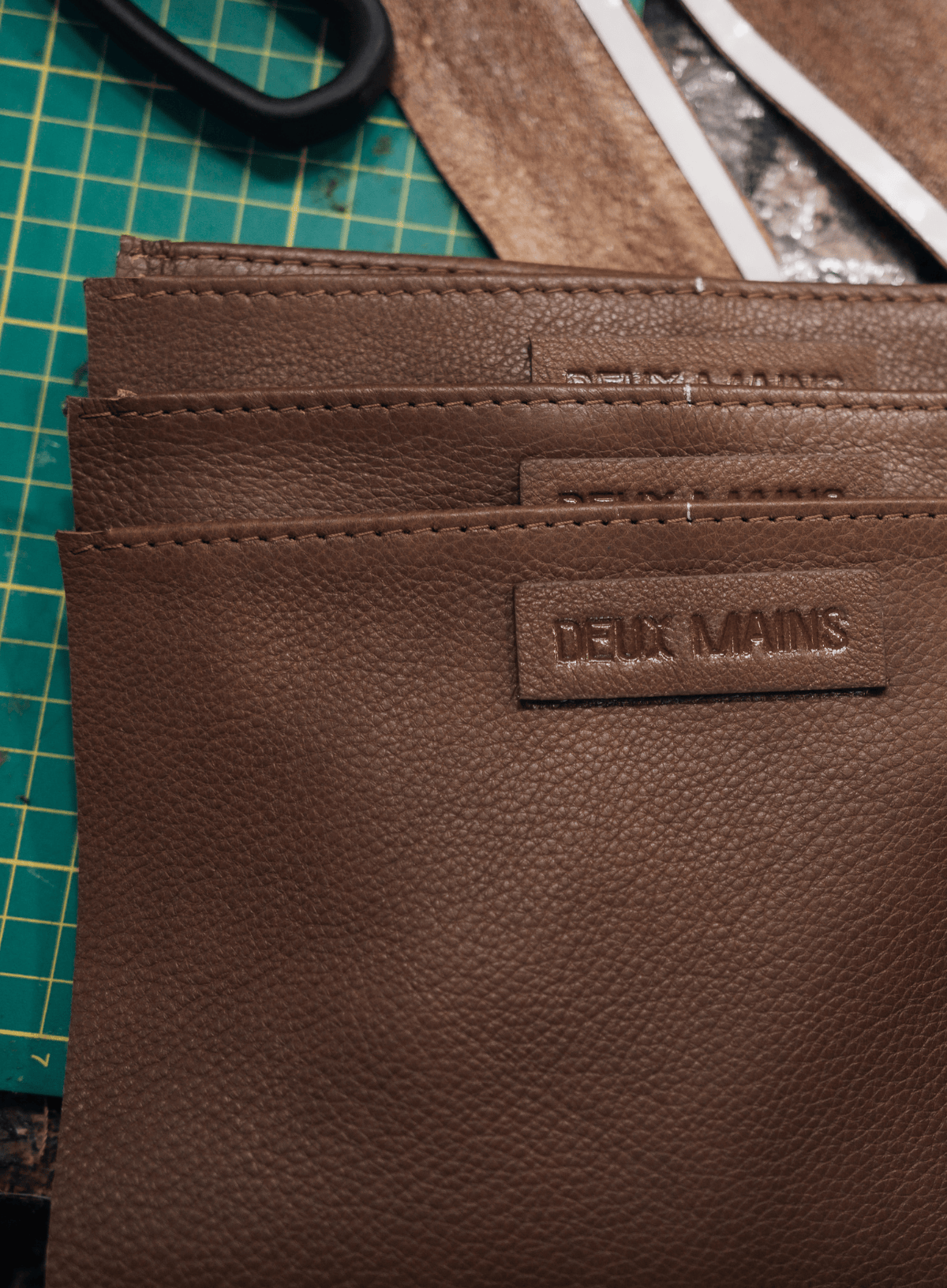 A close-up of a brown textured leather wallet or card holder with a debossed 'DEUX MAINS' logo patch, resting on a green cutting mat with leatherworking tools in the background.