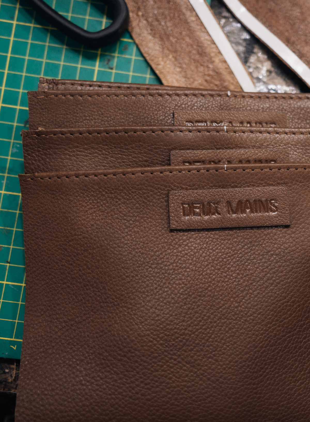 A close-up of a brown textured leather wallet or card holder with a debossed 'DEUX MAINS' logo patch, resting on a green cutting mat with leatherworking tools in the background.