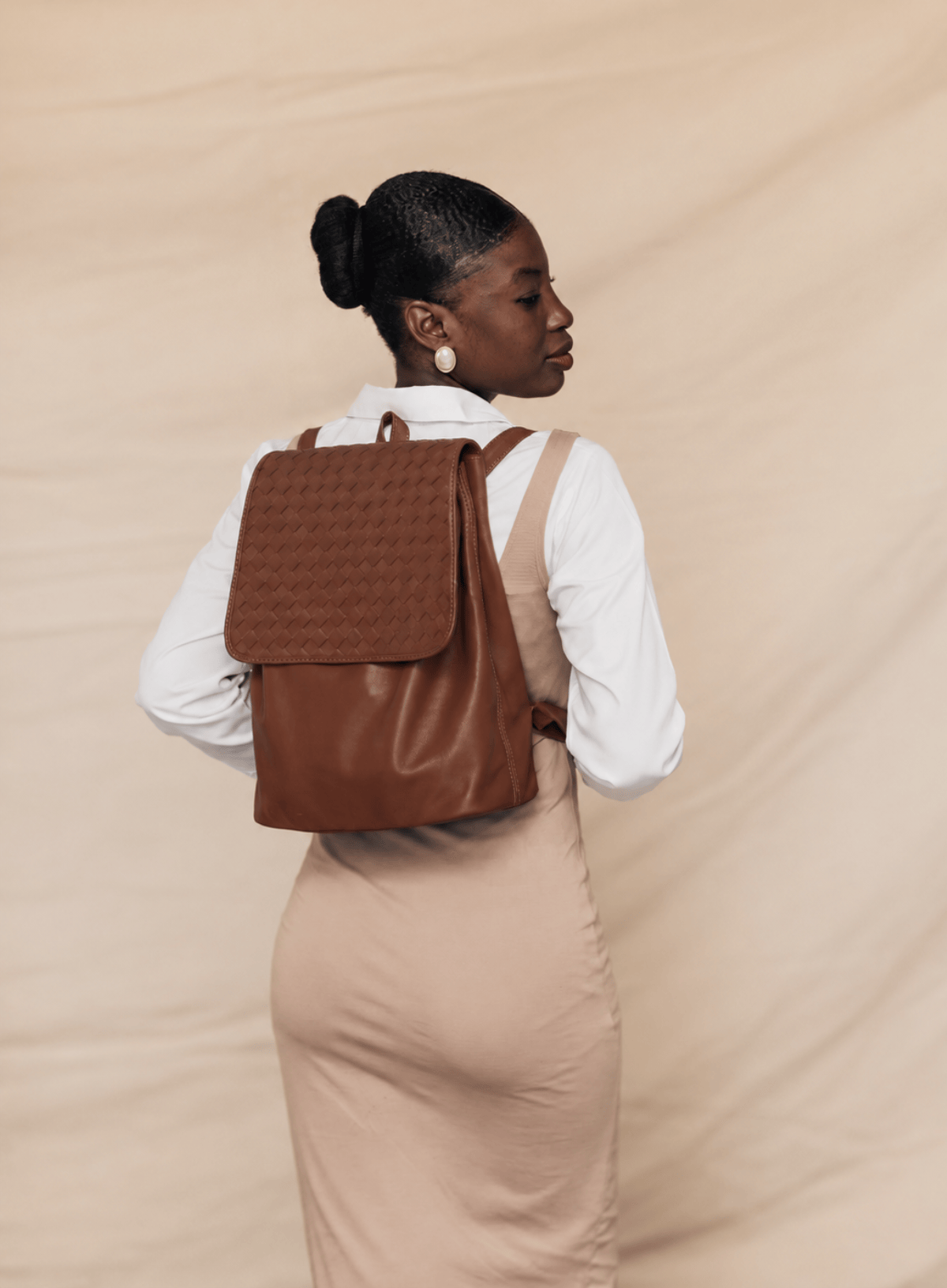 A woman wearing a brown leather backpack with a textured woven flap, styled with a white long-sleeve shirt and a beige dress against a beige fabric backdrop.