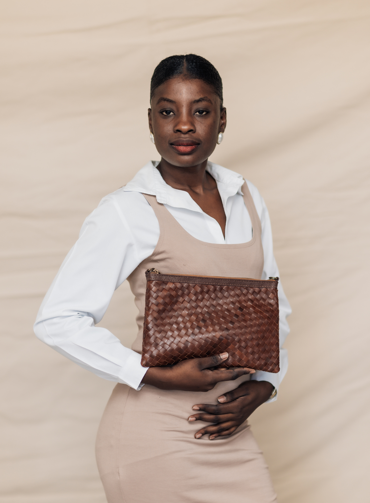 A woman wearing a white collared shirt and a beige dress holds a brown woven leather clutch bag against a beige textured background.
