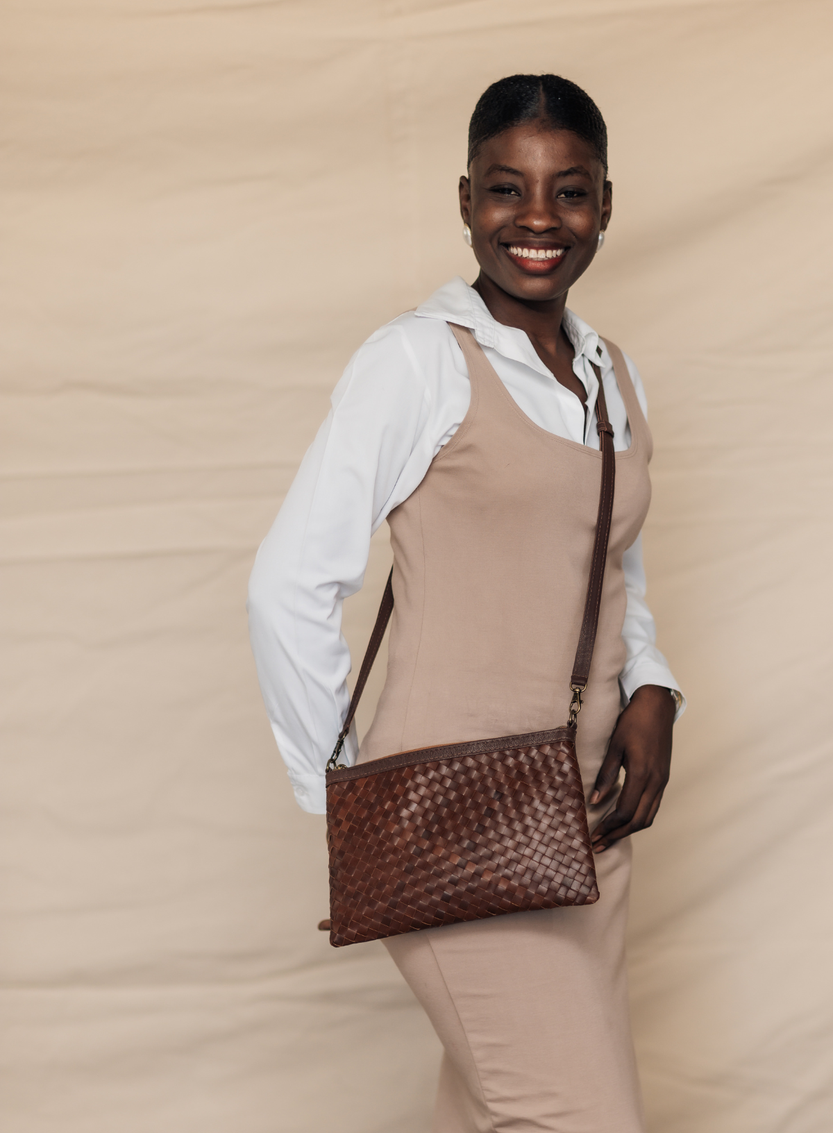 A smiling woman models a brown woven leather crossbody clutch bag with a long strap, worn over a beige dress and white shirt against a beige fabric backdrop.