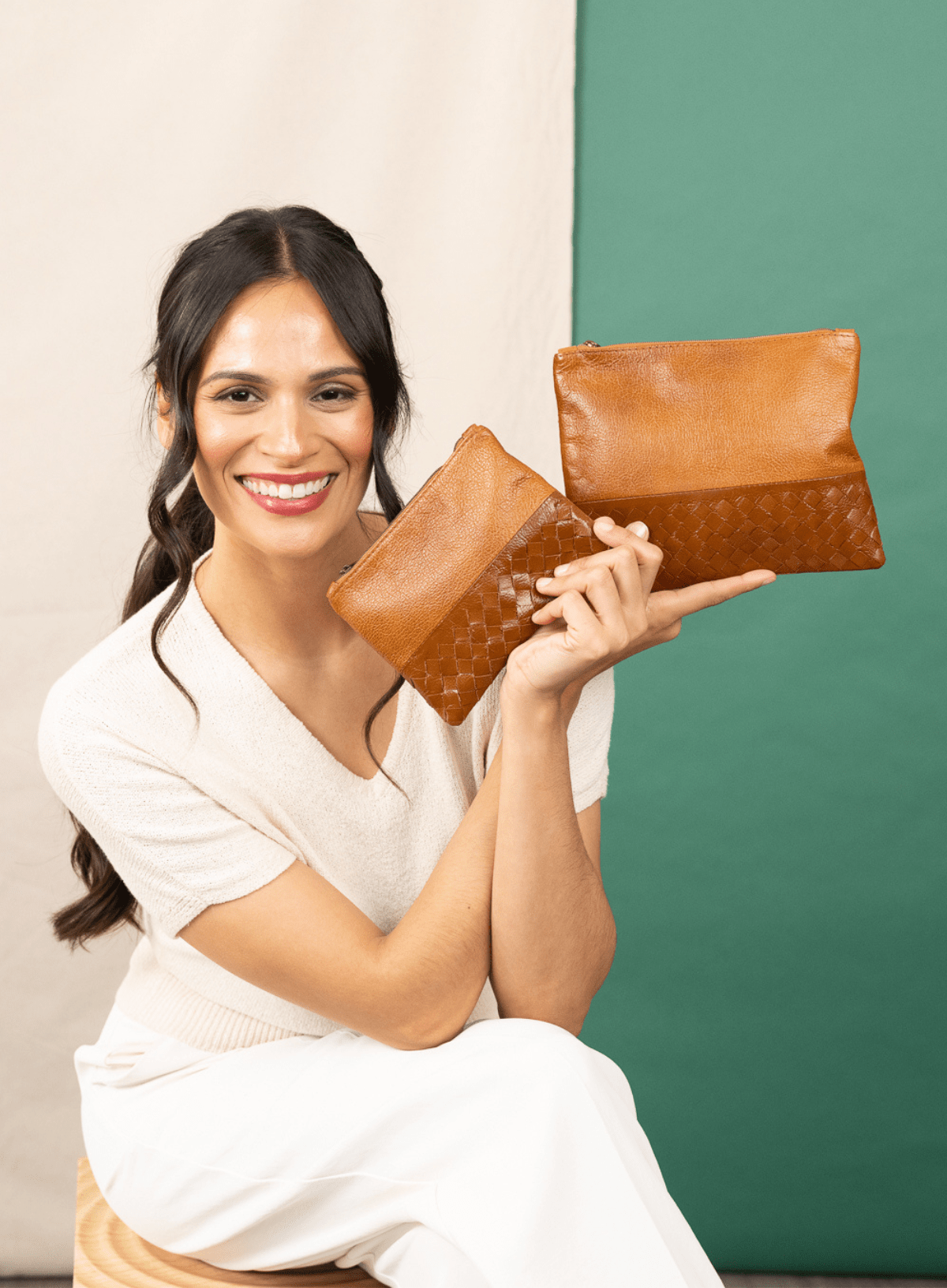 Woman holding two cognac leather pouches against a green and beige background