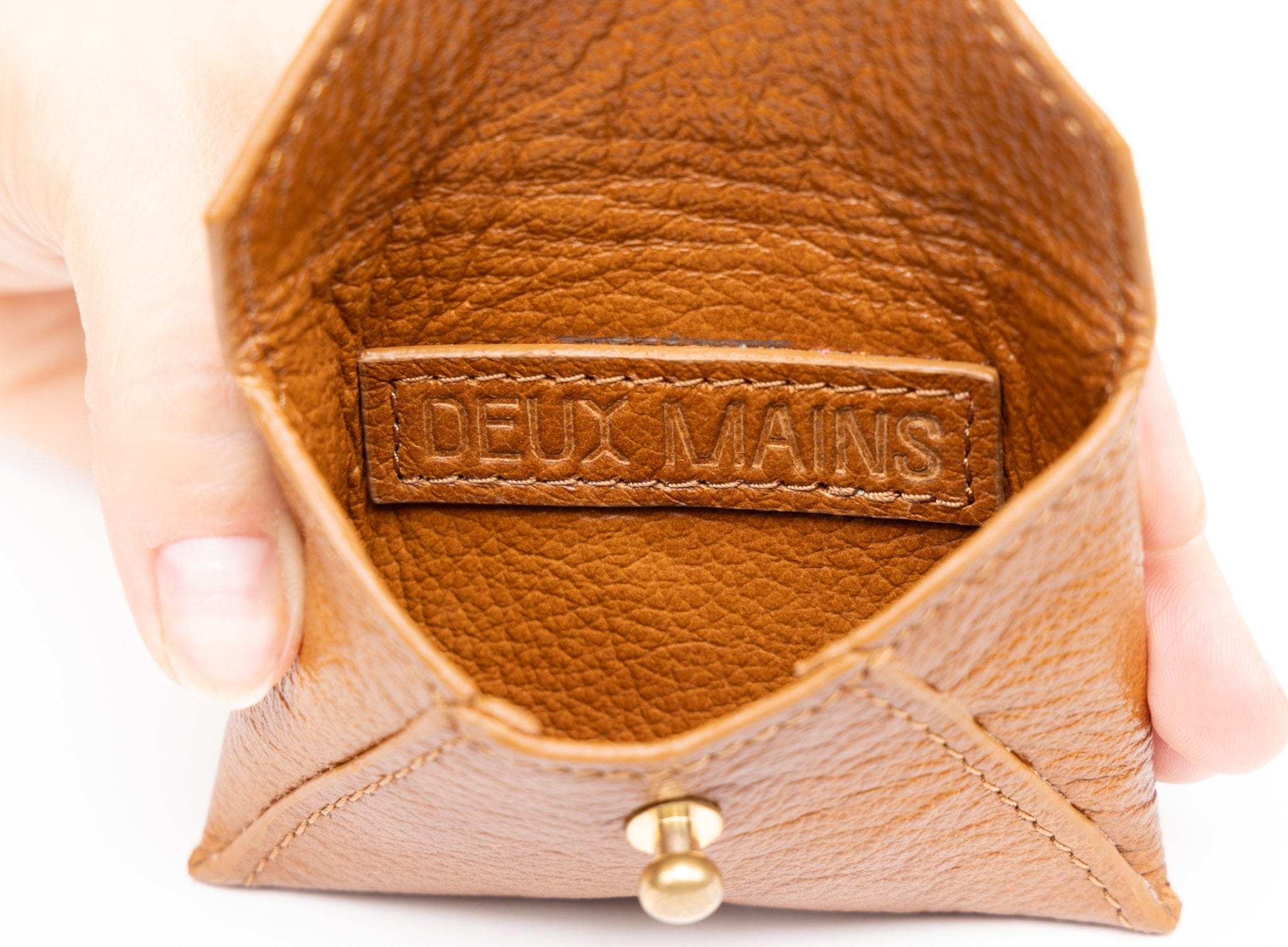 Cognac leather product with 'DEUX MAINS' branding held by a hand on a white background