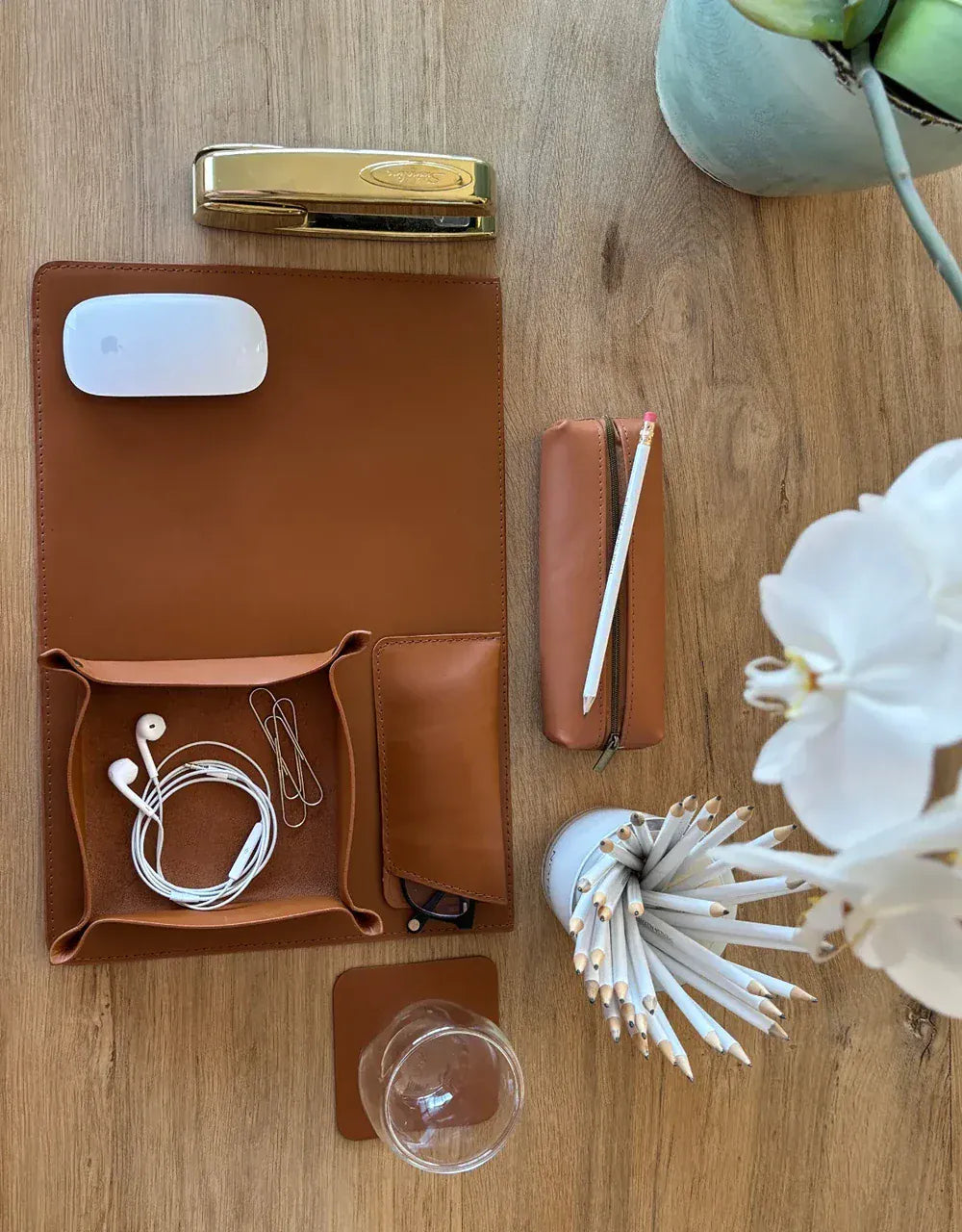 Collection of items including a brown leather case, gold clip, white mouse, and earbuds on a wooden surface.