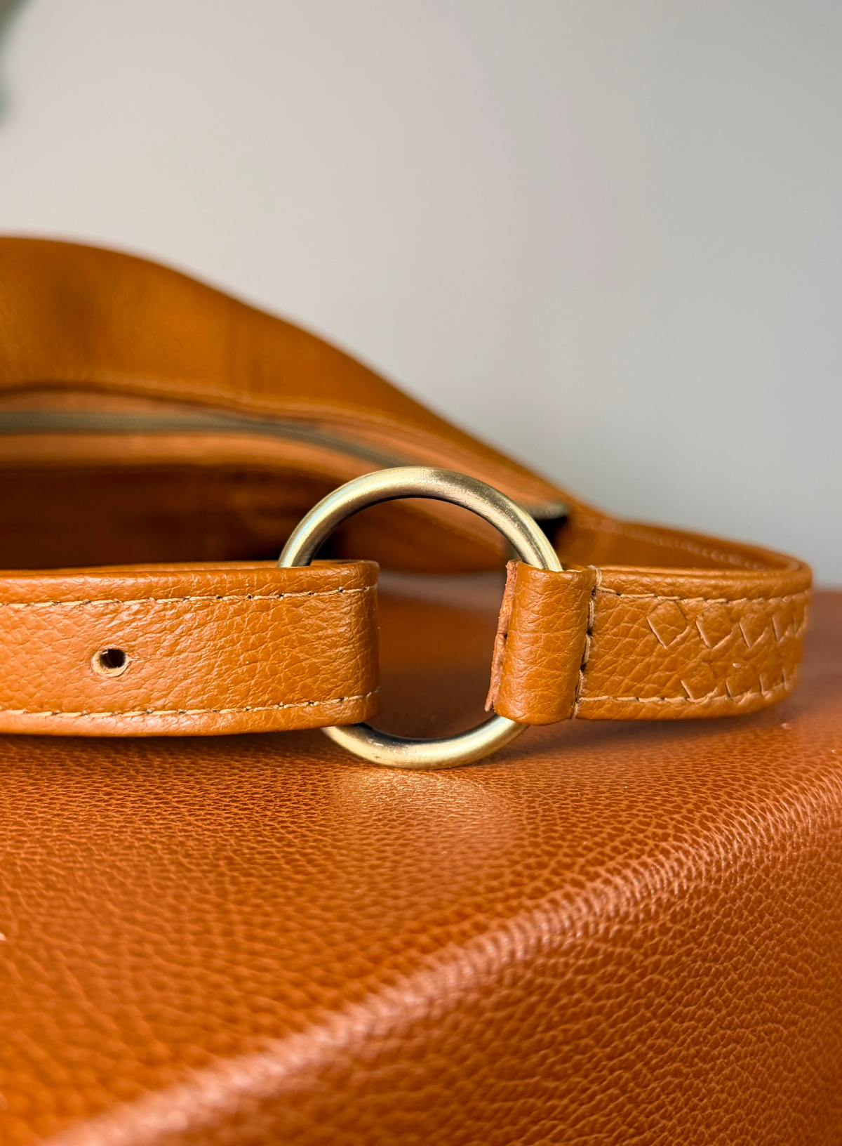 Close-up of a tan leather strap with a large round gold-tone metal ring closure, resting on a textured brown leather surface against a light gray background.
