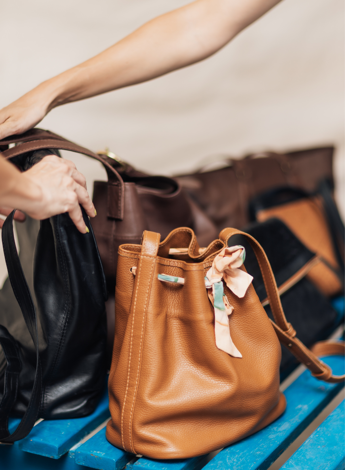 A tan pebbled leather bucket bag with a light pink satin ribbon bow sits on a blue wooden surface, surrounded by other handbags and a person's hands and arm adjusting the bag.