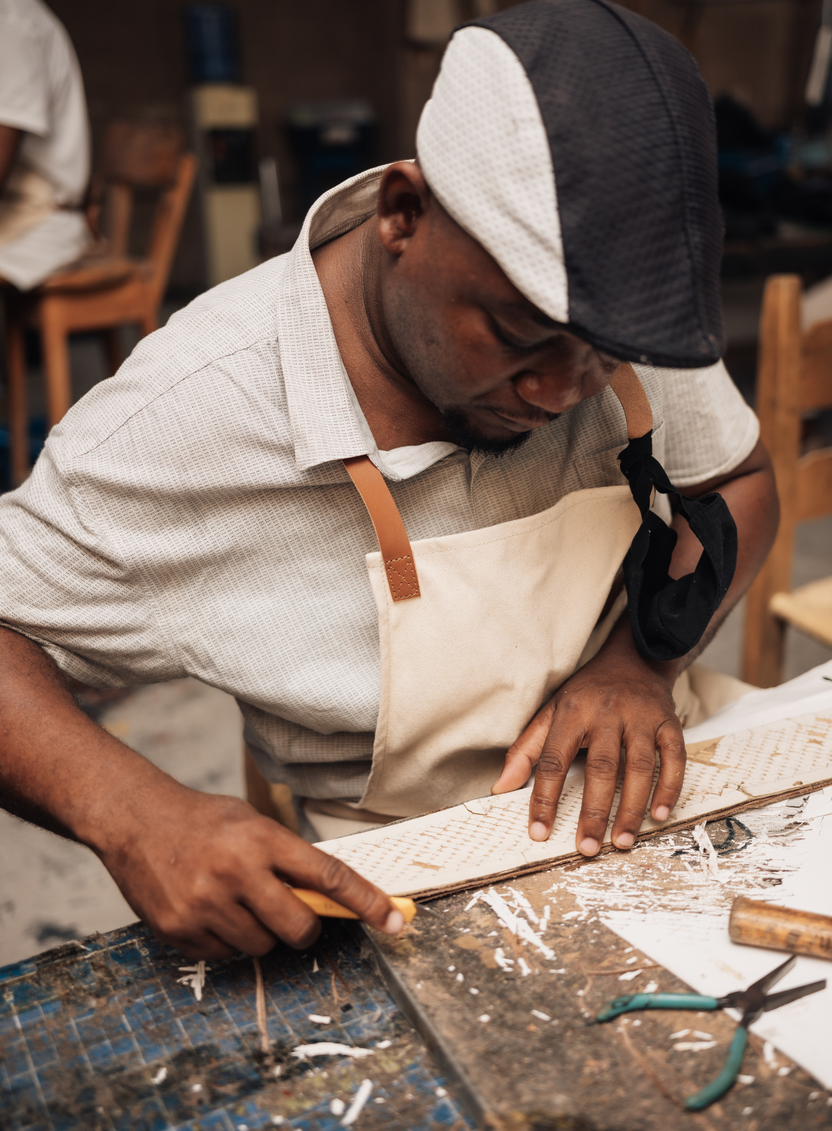 A craftsman wearing a beige apron and a black-and-white cap works at a wooden table, carving or shaping a piece of wood with a chisel. A pair of pliers rests on the workbench nearby.