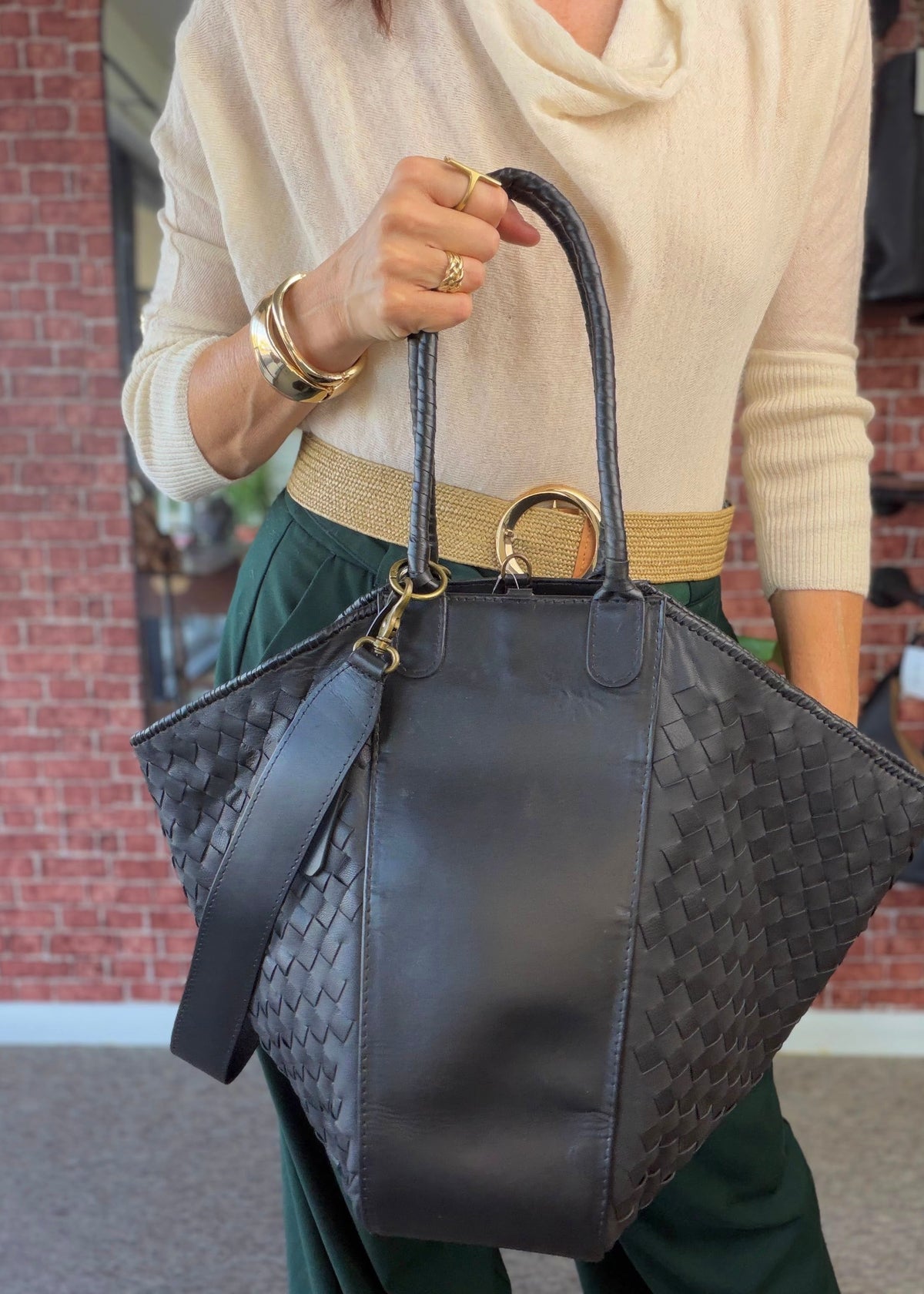 A person wearing a cream cowl-neck sweater and dark green pants holds a large black woven leather tote bag with brass hardware in front of a brick wall.