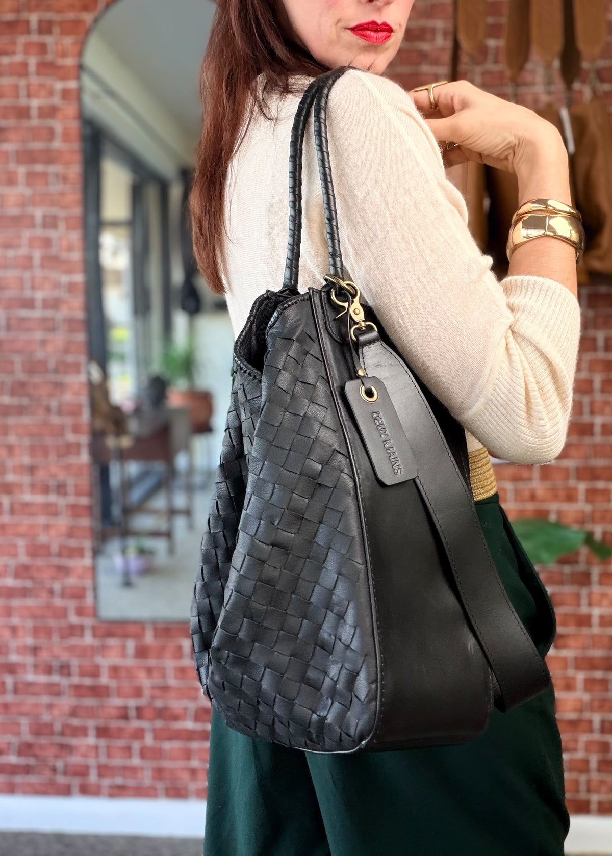 A woman wearing a cream ribbed sweater and dark green skirt carries a black woven leather shoulder bag with a wide strap. The background features a red brick wall and a mirror reflecting the interior of a shop.