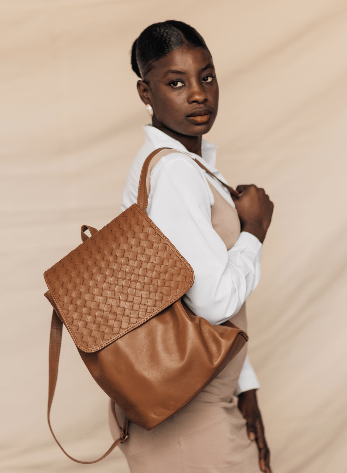 A woman wearing a white collared shirt and beige dress carries a large brown woven leather backpack with a flap cover against a beige backdrop.