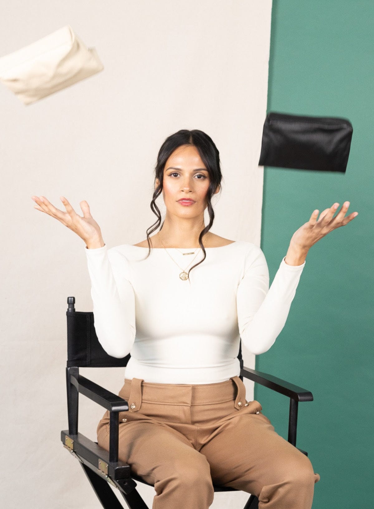 Woman sitting on a director's chair with makeup bags above her head against a white and green background