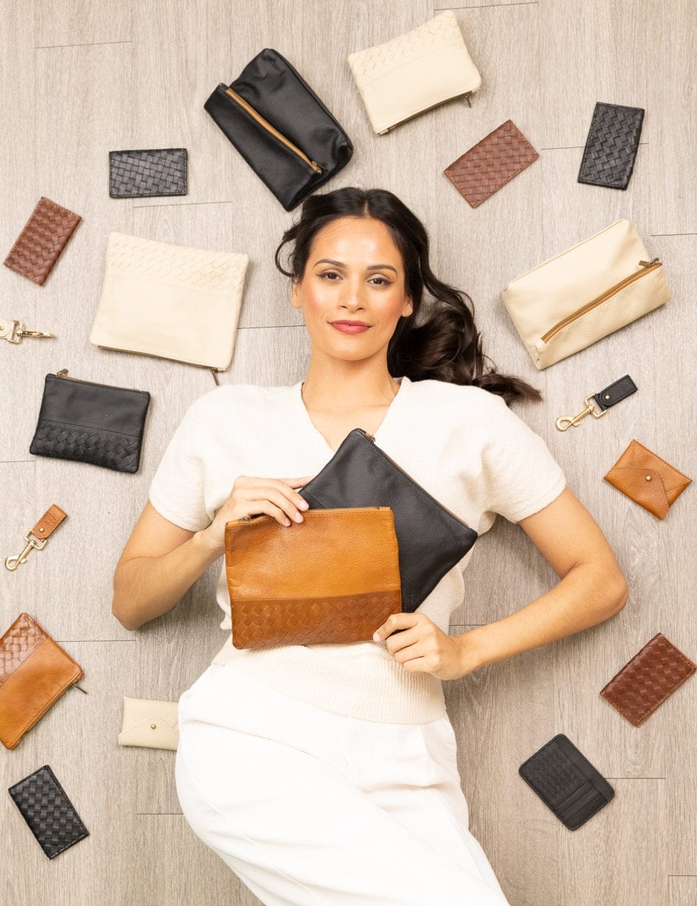 Woman holding a brown and black pouch surrounded by various leather bags on a wooden floor.