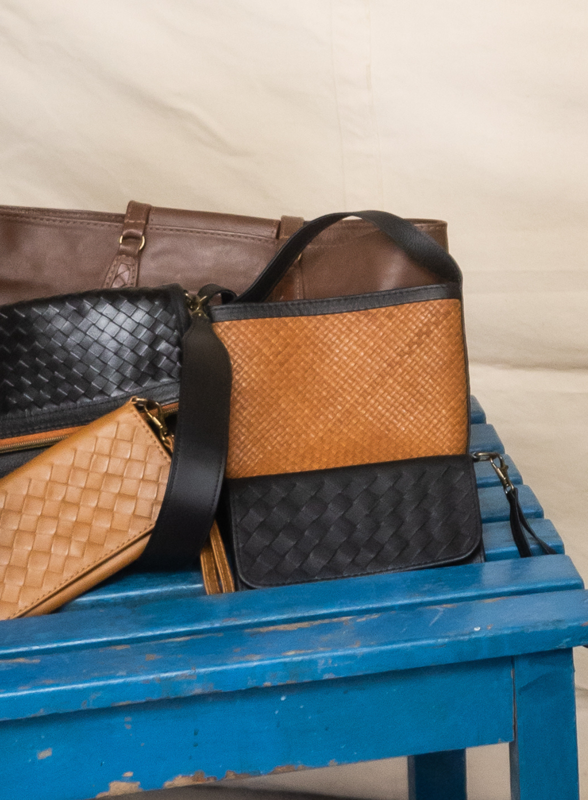 A set of leather handbags and a clutch in black and tan woven and smooth leather, displayed on a distressed blue wooden bench against a cream fabric backdrop.