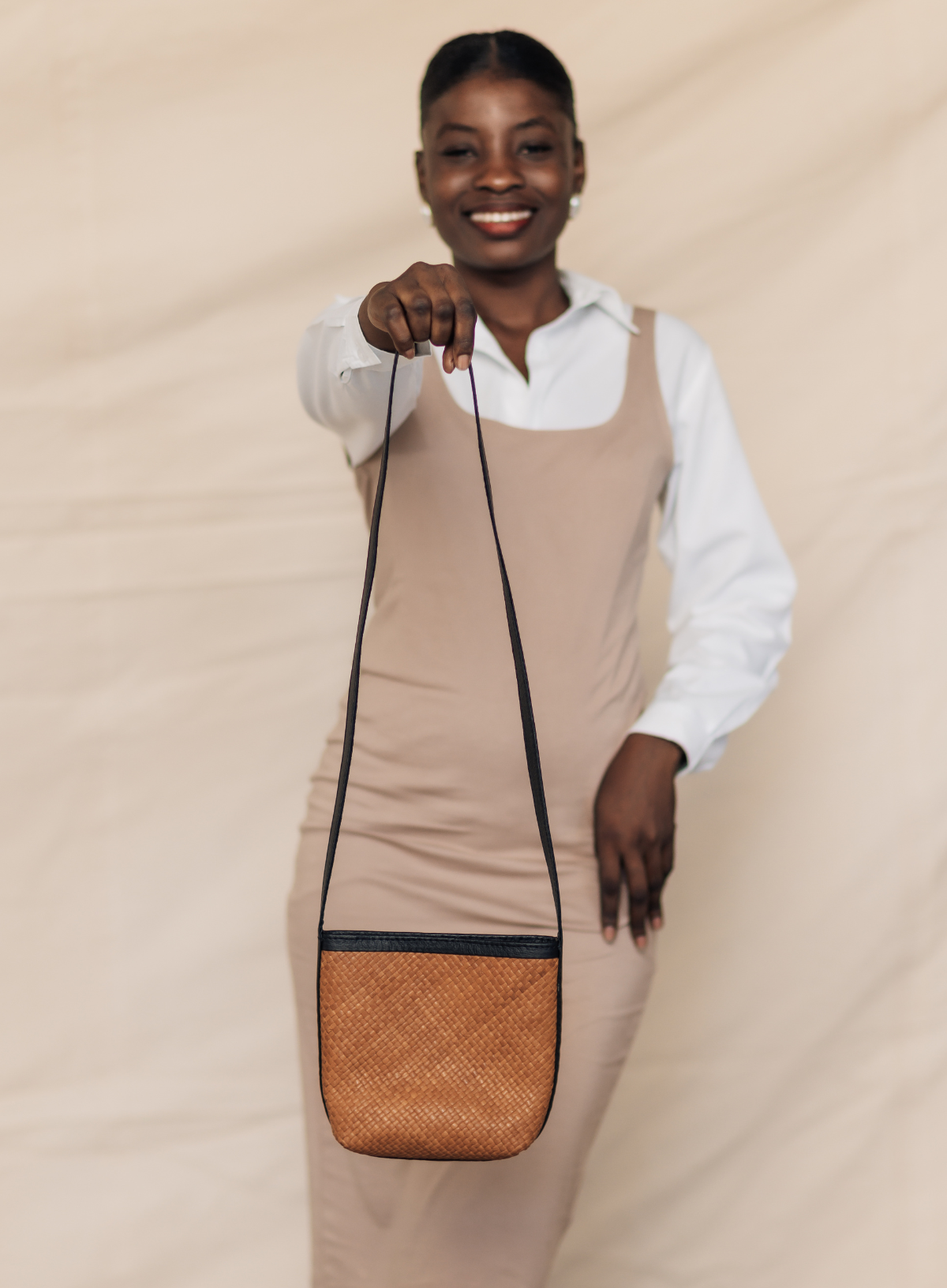A smiling woman wearing a beige pinafore dress over a white collared shirt holds up a small crossbody bag with a woven tan body and black strap against a draped beige backdrop.
