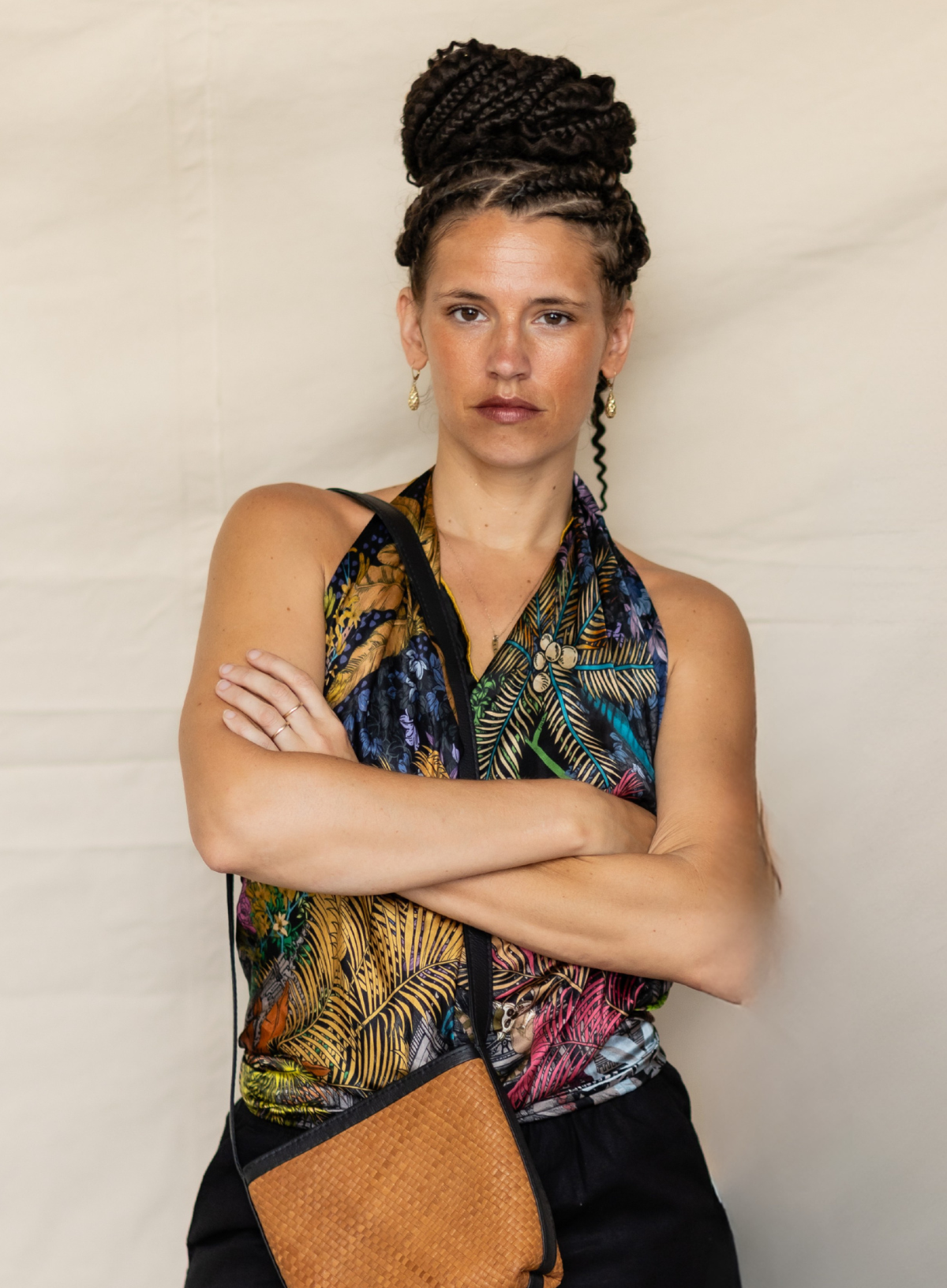 A woman with braided hair in a bun wearing a colorful tropical print halter top and a woven brown crossbody bag, posing with her arms crossed against a light beige backdrop.