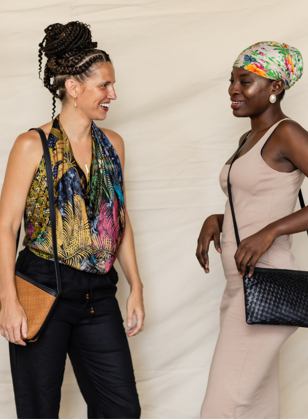 Two women standing and smiling at each other against a cream fabric backdrop. The woman on the left wears a colorful tropical print halter top, black pants, and a woven brown shoulder bag.