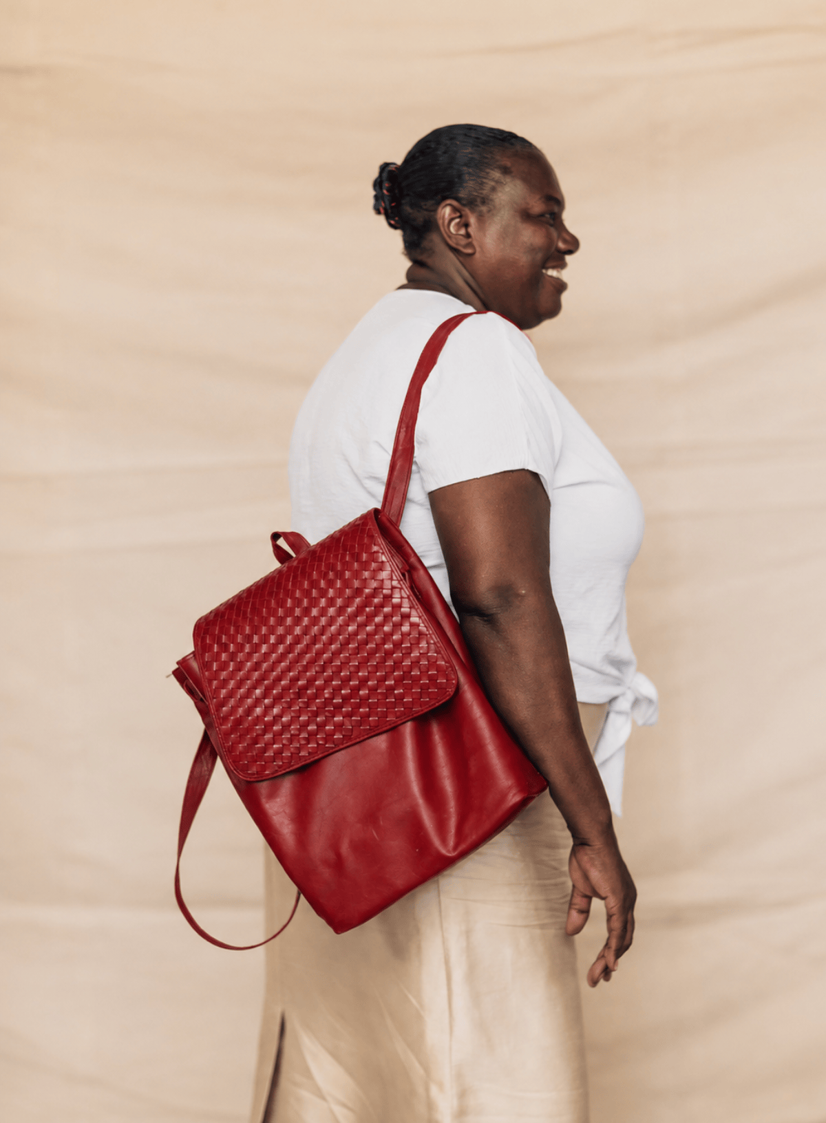 A woman in a white t-shirt and beige skirt is shown from the back and side profile, carrying a red woven leather backpack with a shoulder strap against a beige fabric backdrop.
