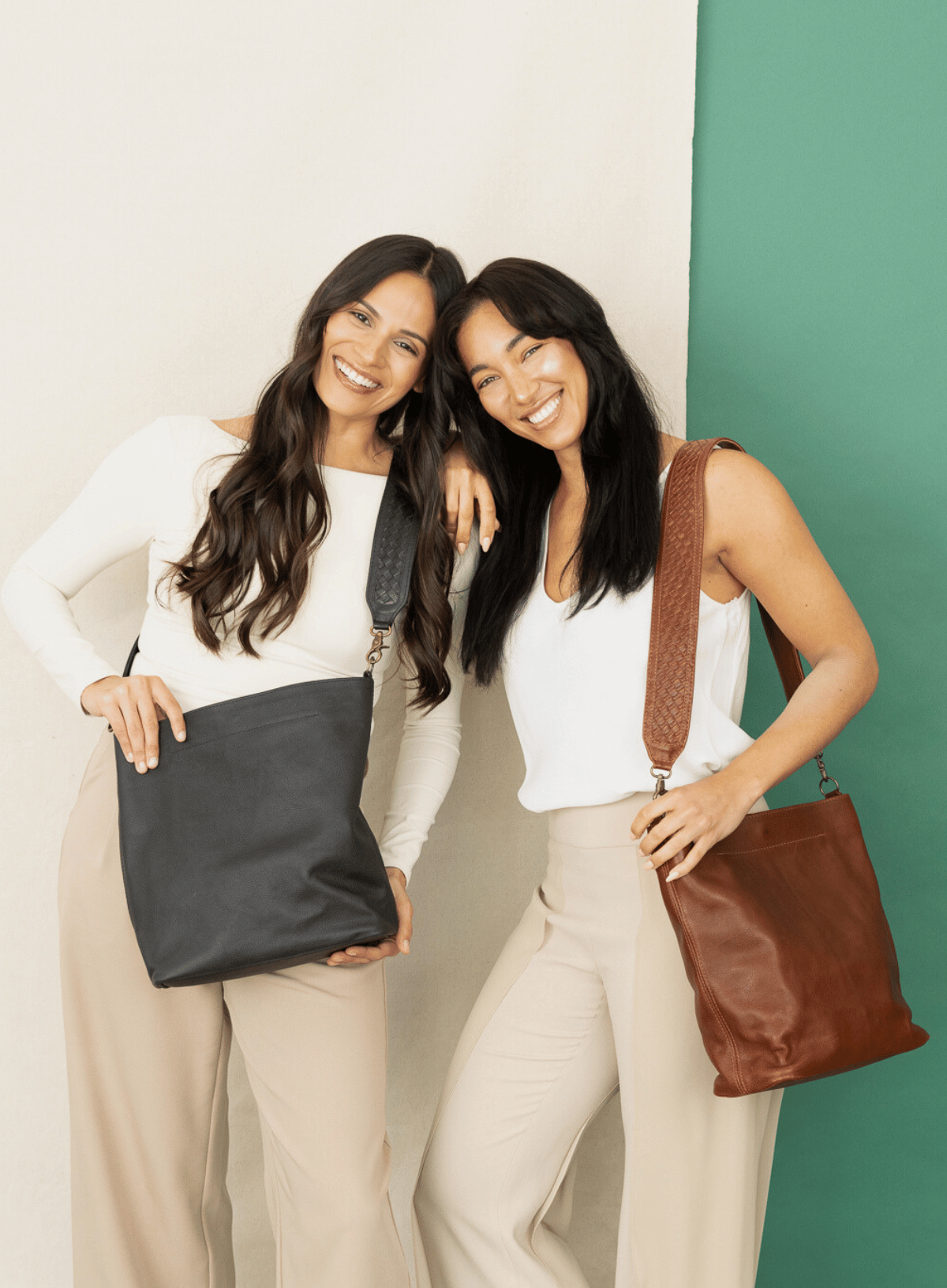 women leaning on each other holding leather totes in front of beige and green background