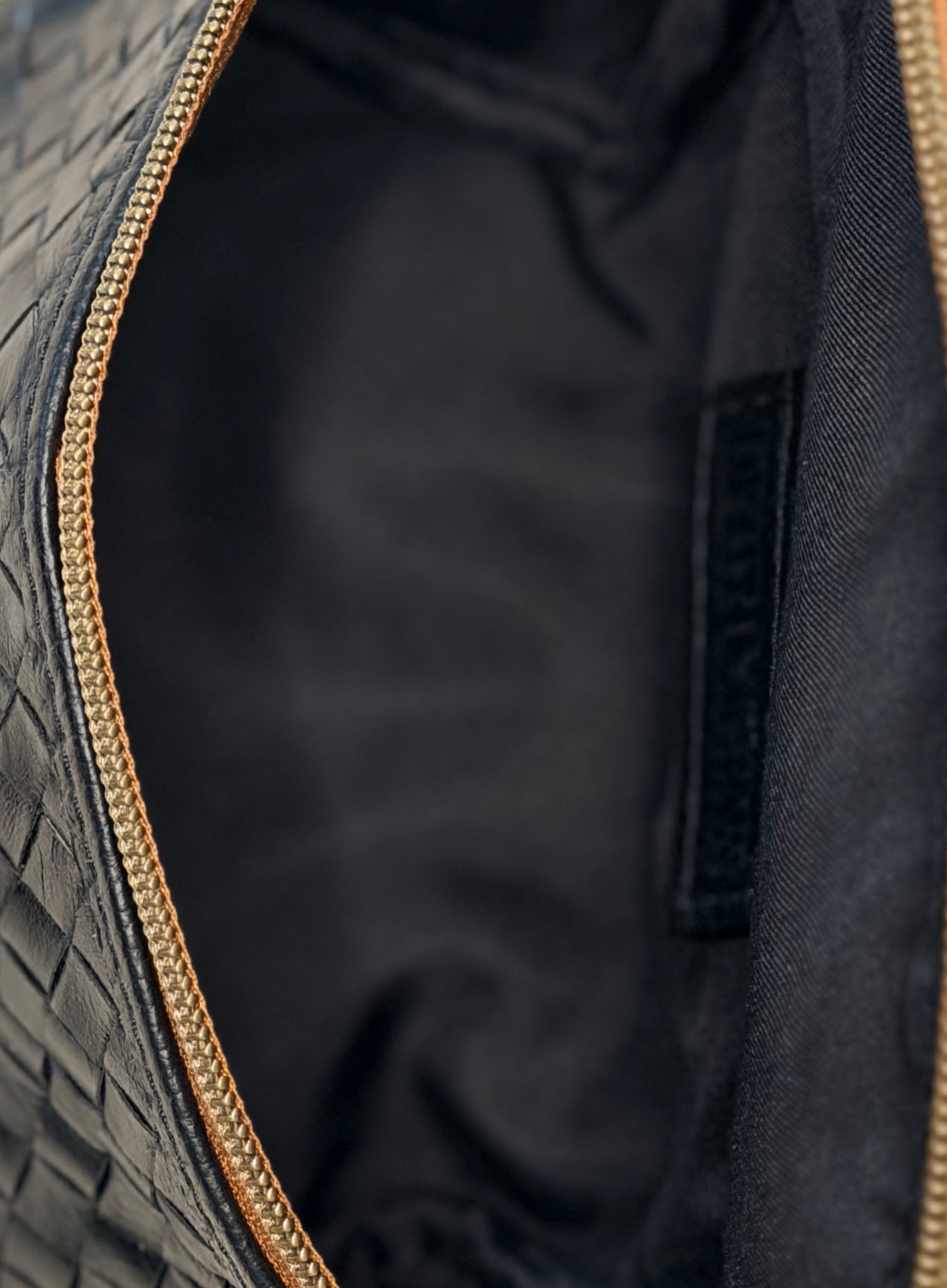 Close-up of the interior of a black woven leather bag, showing a black satin lining and a gold-tone zipper.