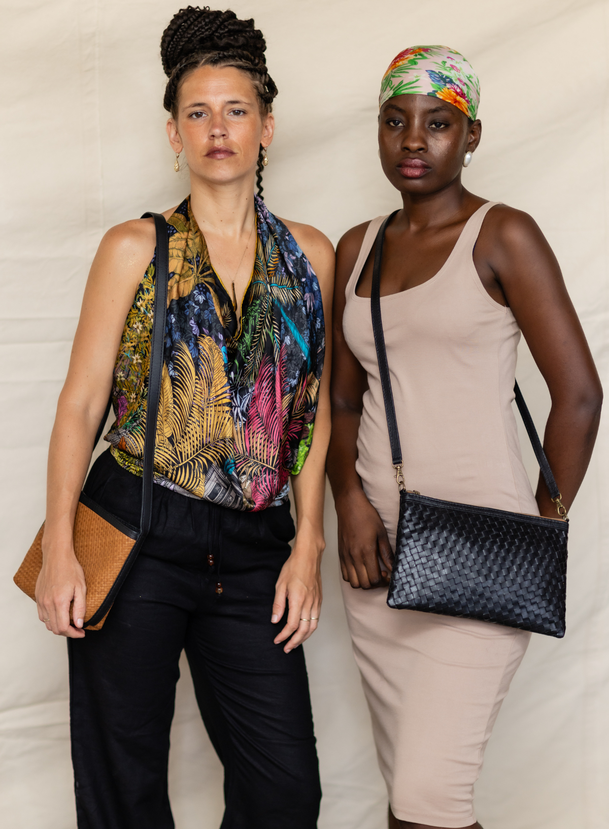 Two models standing side-by-side wearing crossbody bags. The model on the left wears a colorful tropical print halter top and black pants, holding a brown leather clutch.