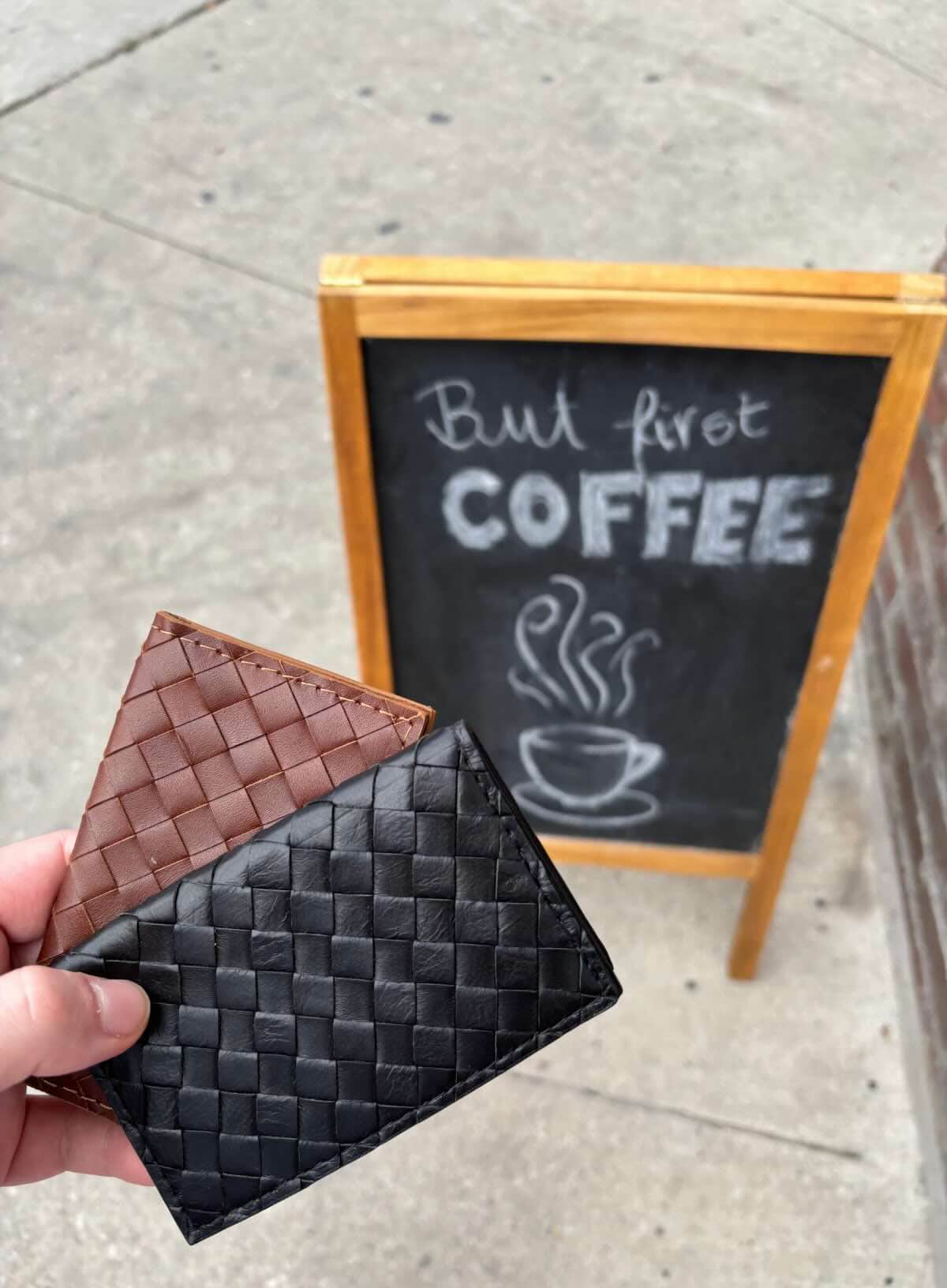 Two woven leather wallets, one brown and one black, held in front of a chalkboard sign on a sidewalk.