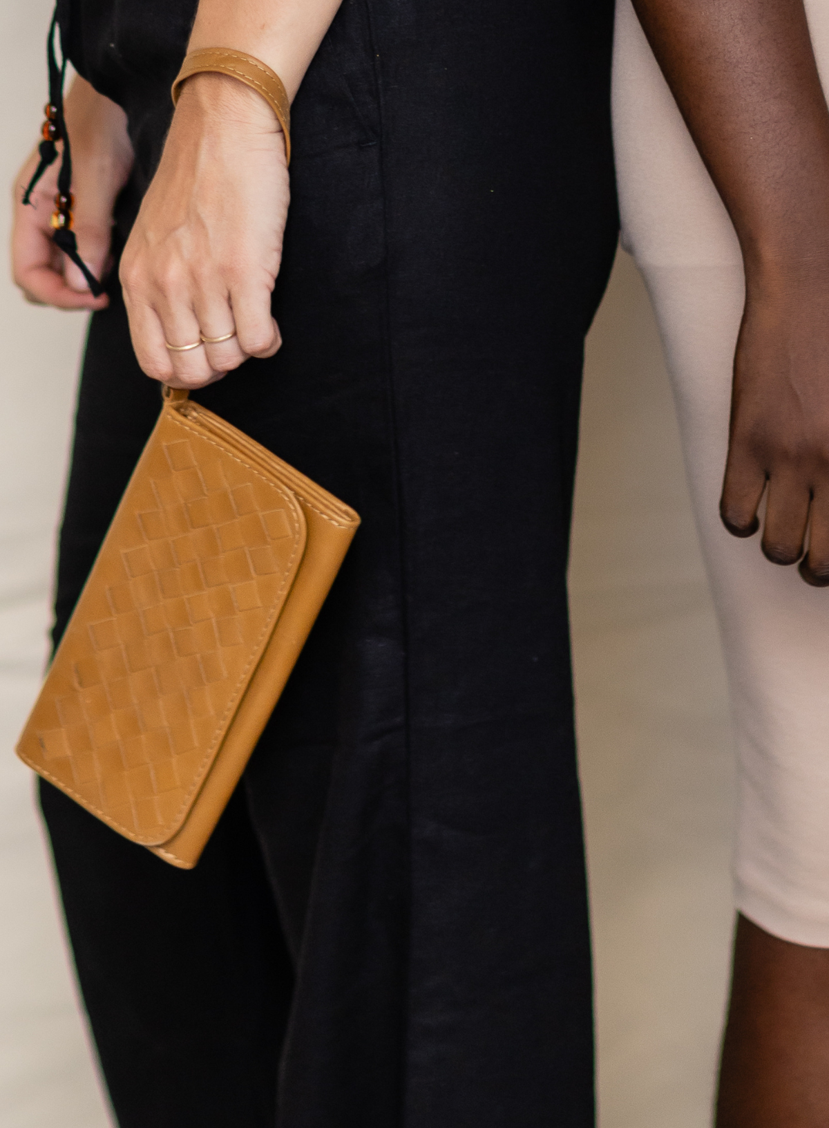 A close-up lifestyle shot of a person holding a tan woven leather clutch bag, wearing a black dress and a gold bangle.