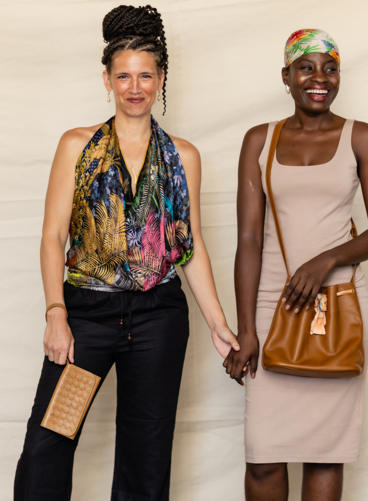 Two women standing side by side holding hands, wearing colorful tropical-print clothing and accessories against a light beige backdrop. The woman on the left wears a multicolor scarf, black pants, and a tan clutch.