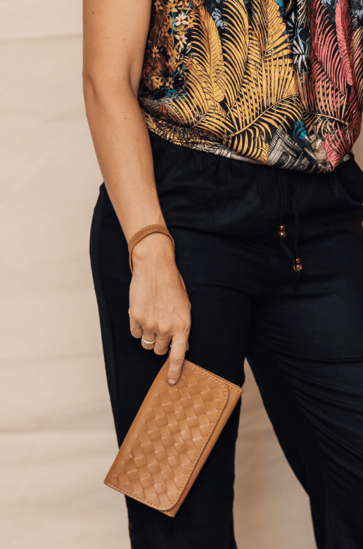 A lifestyle shot of a woman wearing a tropical print top and black pants, holding a brown woven clutch. She is standing next to another person in a beige dress holding a brown leather shoulder bag, against a neutral beige backdrop.