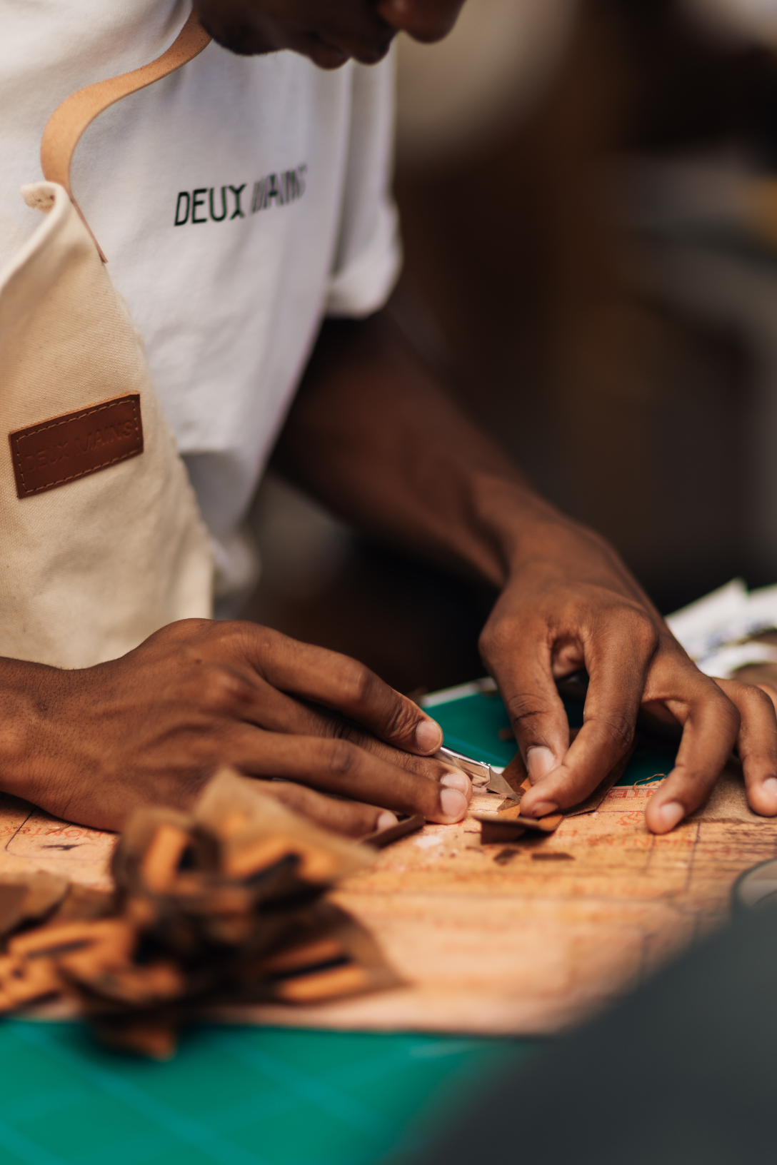 A close-up lifestyle shot of a leather craftsman's hands working on a leather piece over a green cutting mat, with the artisan wearing a white t-shirt and beige apron.