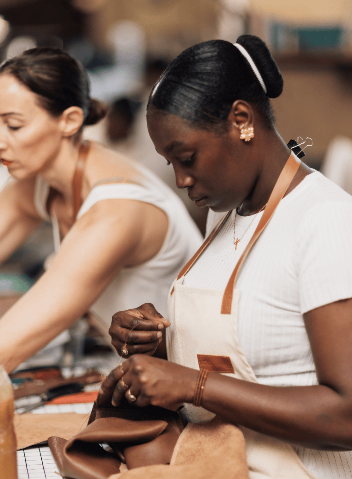 A woman in a white apron works at a leathercraft table, shaping a brown leather piece, with another woman working in the background in a warm, softly blurred workshop setting.