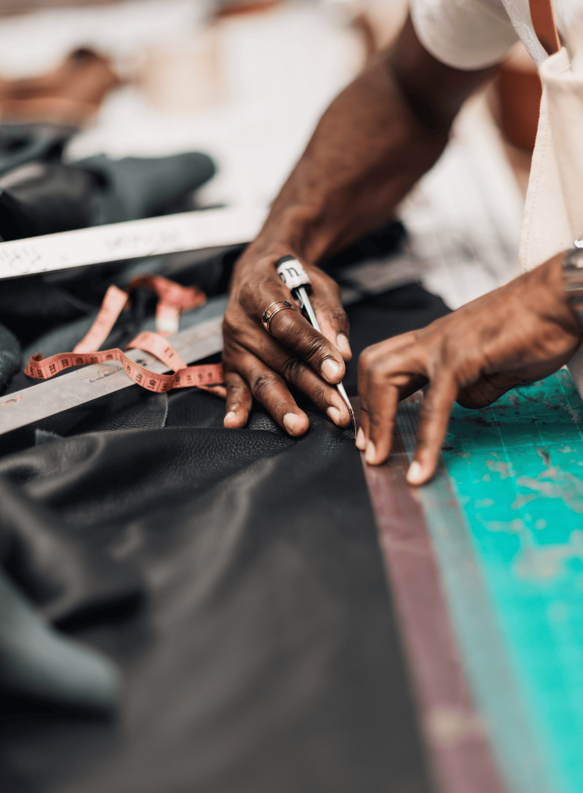 Close-up of a tailor's hands marking black fabric with a pencil, surrounded by measuring tools like a tape measure and ruler on a worktable.