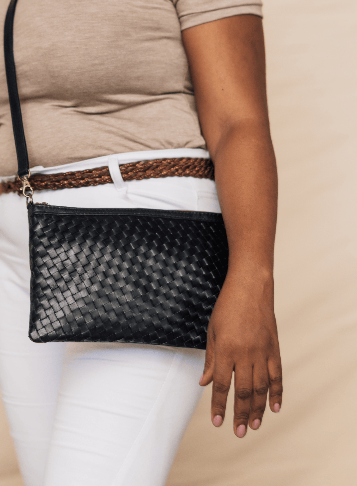 A black woven leather clutch bag with a gold-tone zipper and a thin black shoulder strap, worn crossbody by a model wearing a beige top and white pants.