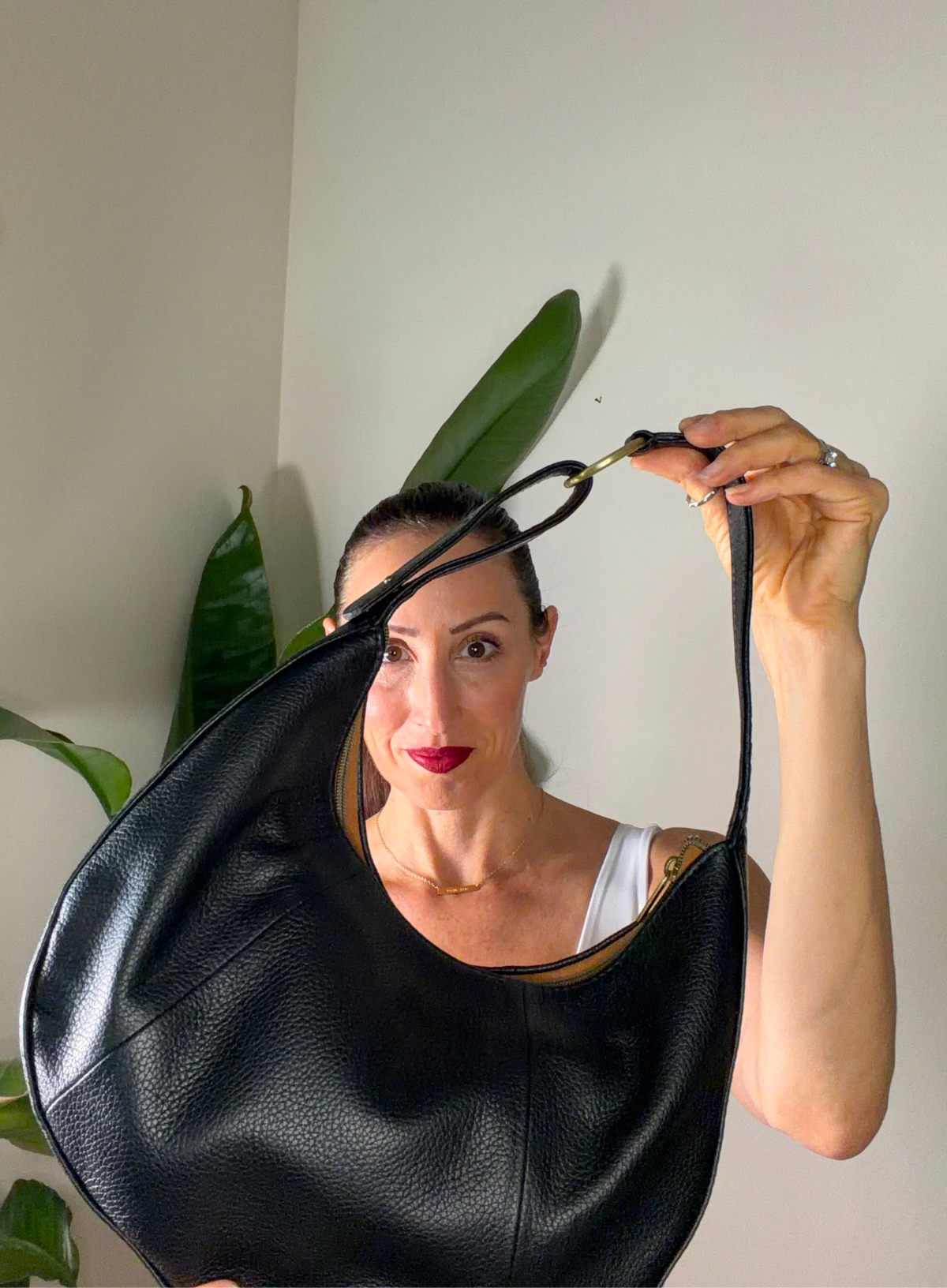 A woman holding up a large black pebbled leather crescent shoulder bag with gold-tone hardware, set against a neutral indoor background with a green plant.