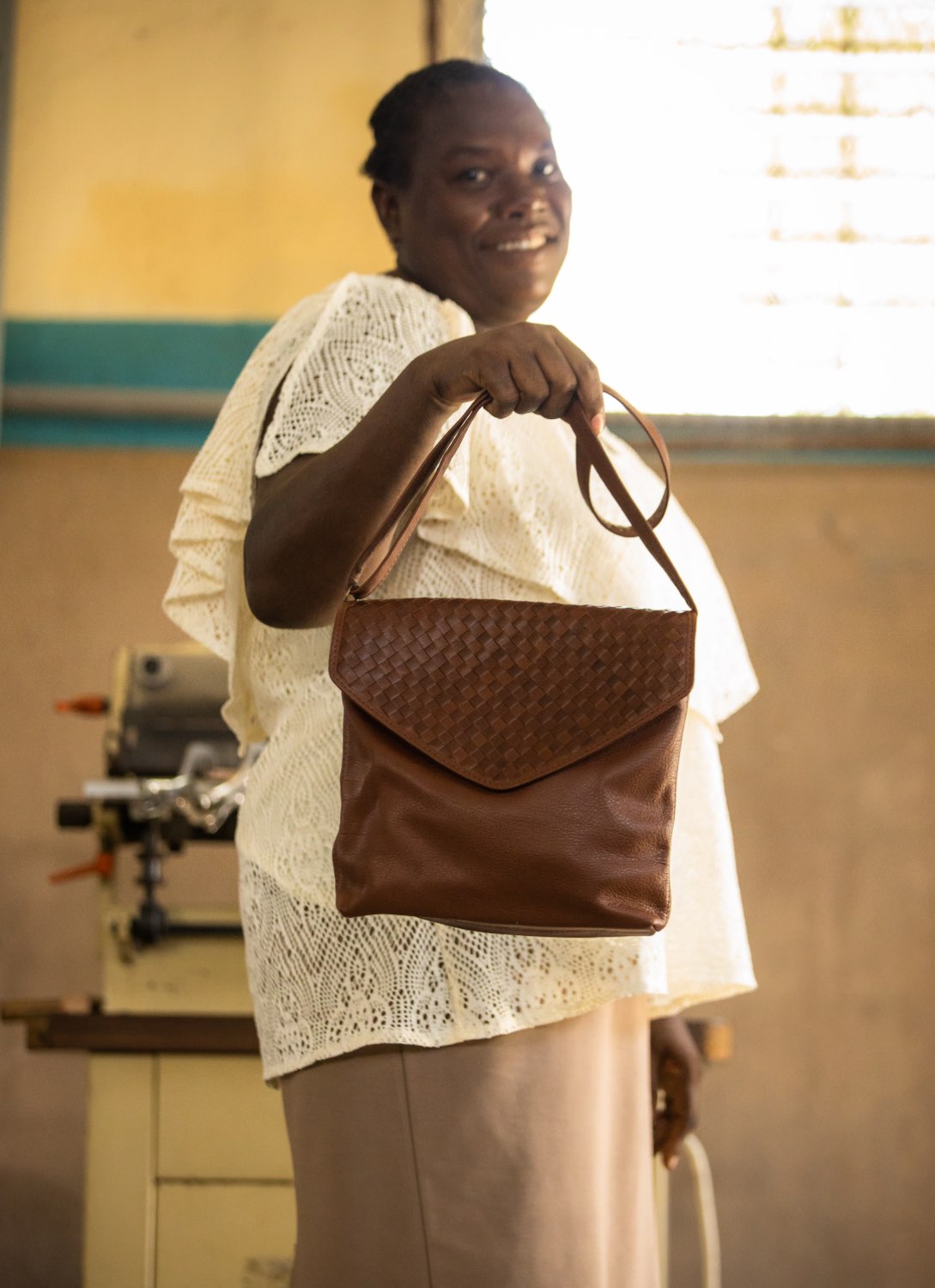 envelope crossbody in brown from spring and summer collection held by haitian artisan in front of haiti factory.