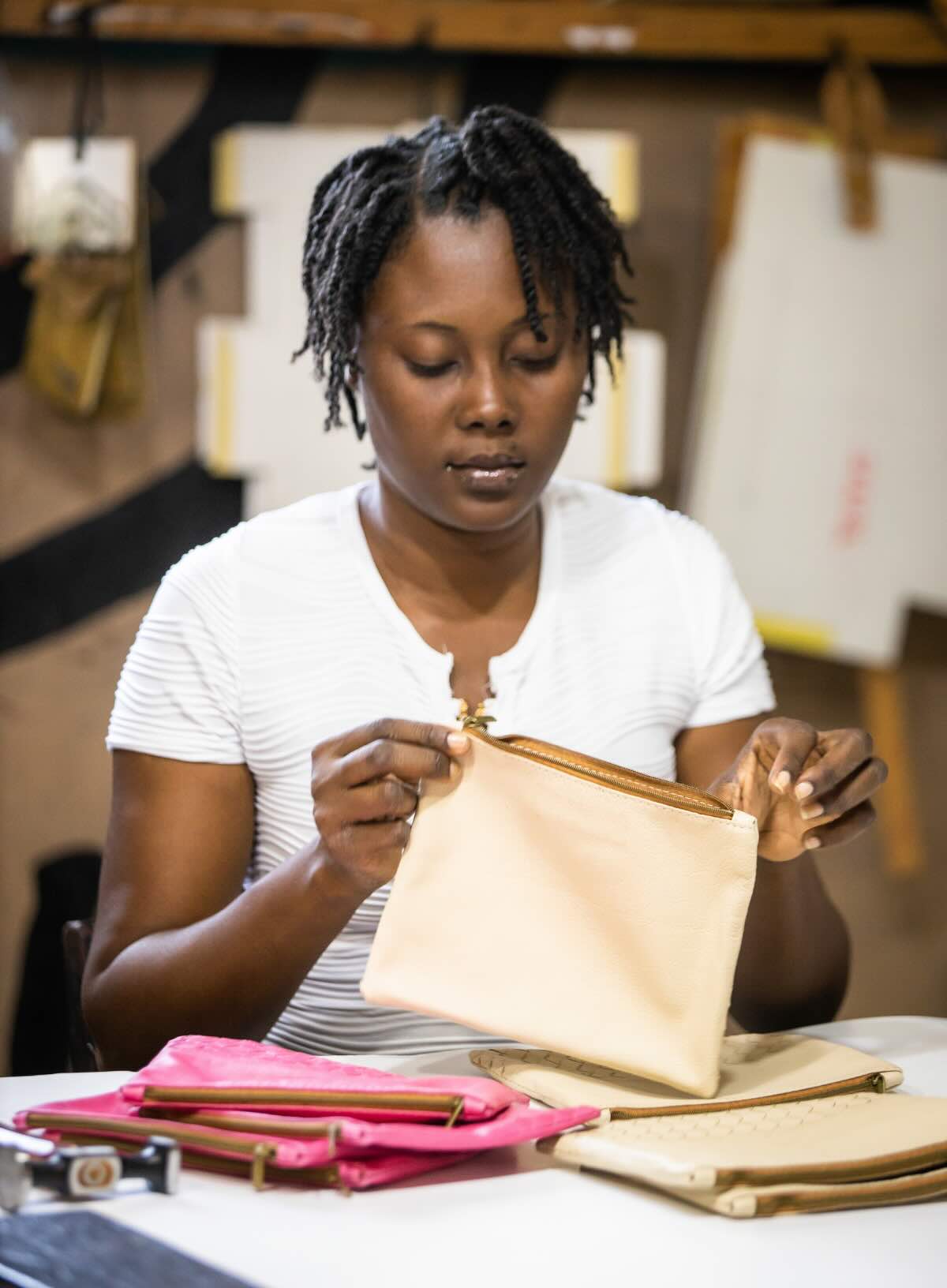 large pouches from ethically crafted accessories in bone and pink next to haitian artisan in factory.