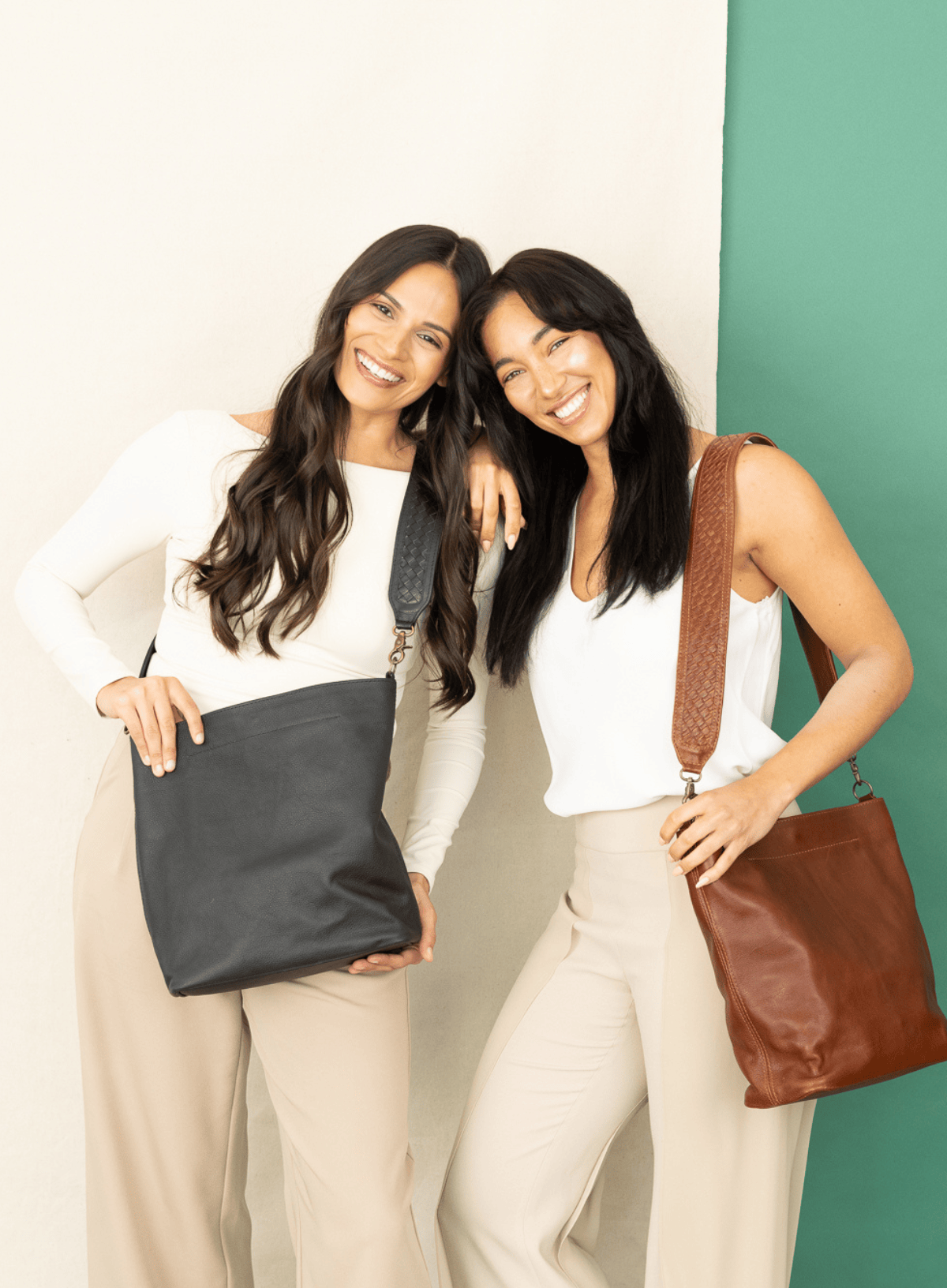Two women posing with leather shoulder bags. The woman on the left holds a large black leather tote bag, and the woman on the right wears a brown leather shoulder bag with a woven strap.