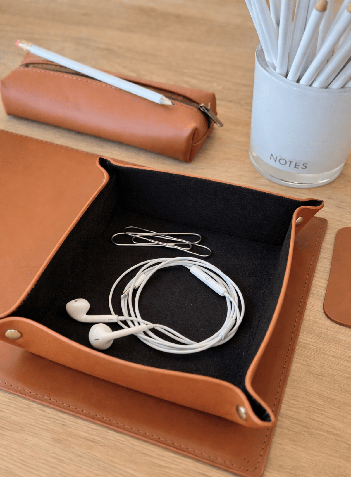 Cognac leather tray with earbuds, glasses case, and stationery on a wooden surface