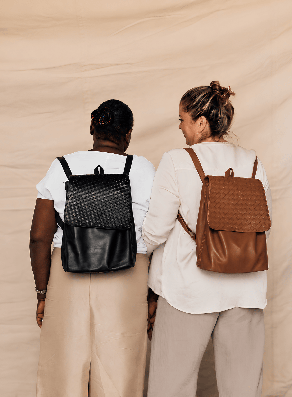 Two women seen from behind wearing woven leather backpacks, one black and one brown, against a beige fabric backdrop.