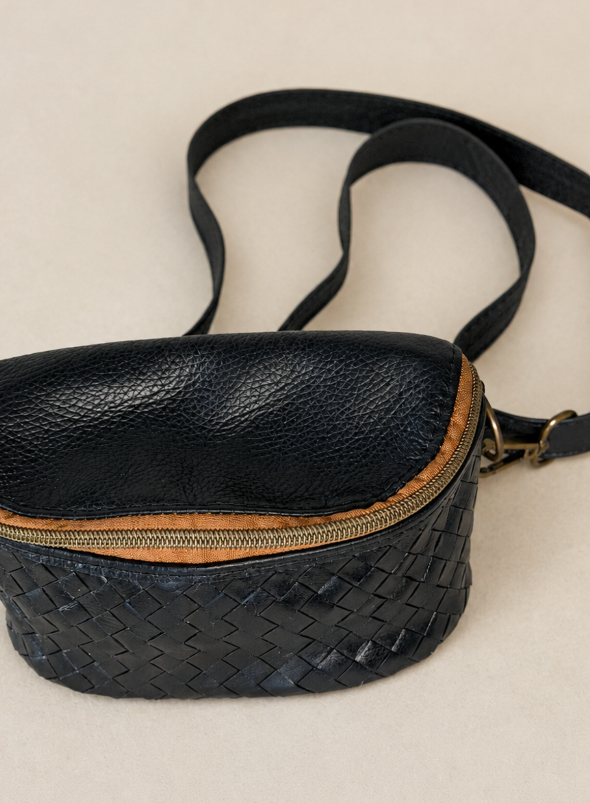 A black leather crossbody bag with a textured pebbled top half, a woven basketweave bottom half, a tan zipper trim, and a black strap, shown on a light beige background.